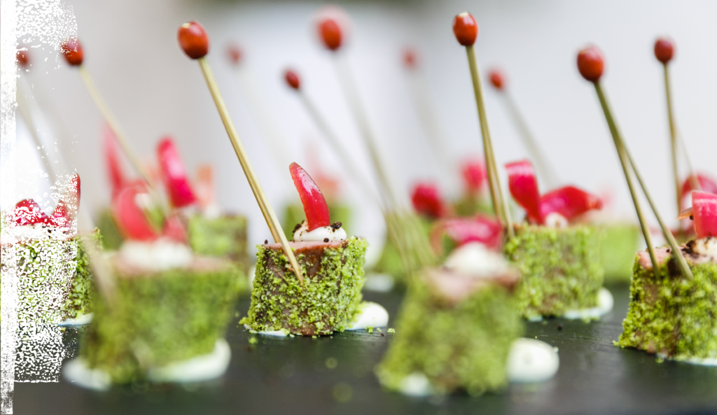 Nahaufnahme von Gourmet-Häppchen mit grünem Kräutermantel und Spießchen, garniert mit rotem Gemüse auf einem Tablett.