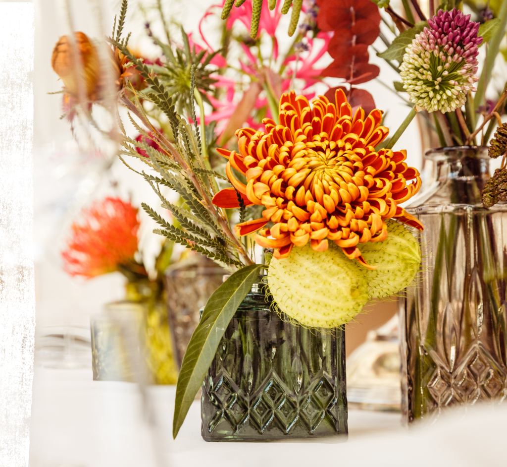 Eine Nahaufnahme von leuchtend orangefarbenen und gelben Blumen in einer dekorativen Glasvase, die jedem Tisch Eleganz verleihen - perfekt für professionelles Catering für Events in Köln, mit weiteren sanft blühenden Blumen im Hintergrund.