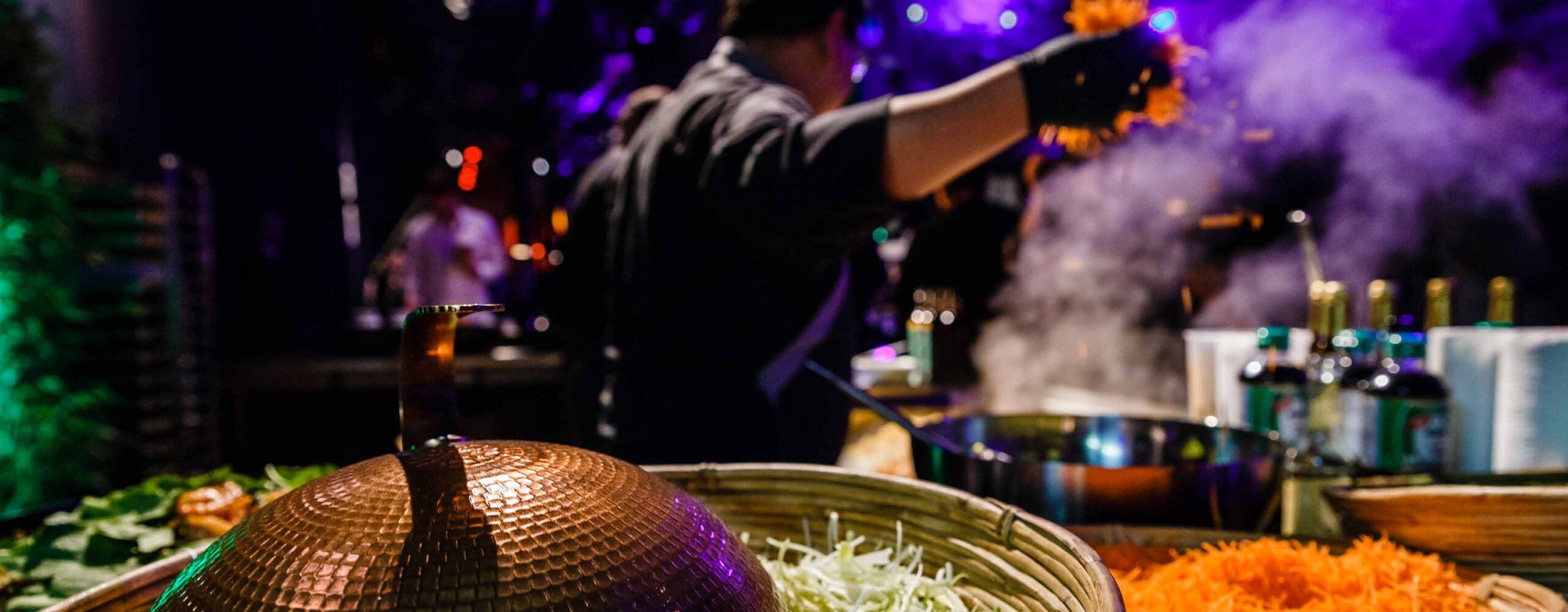 Ein Koch, der an einer Kochstation mit Dampf und bunten Zutaten in einer lebendigen, schummrigen Umgebung kocht - perfekt für Eventcatering für besondere Anlässe.