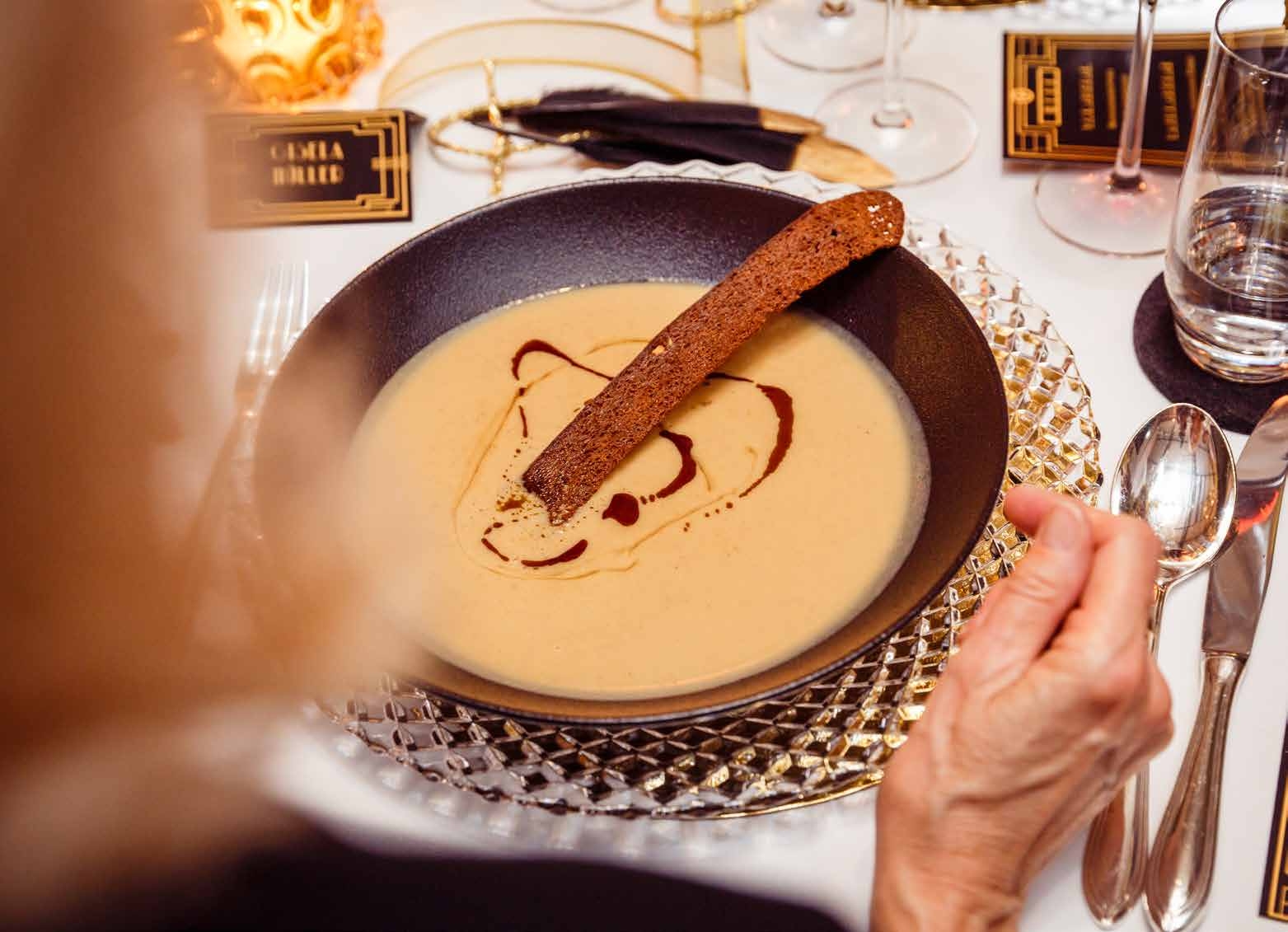 Eine Schale mit cremiger Suppe mit einem Strudelmuster und einer Brotstange obenauf, auf einem dekorativen Tisch - perfekt für Eventcatering für besondere Anlässe.