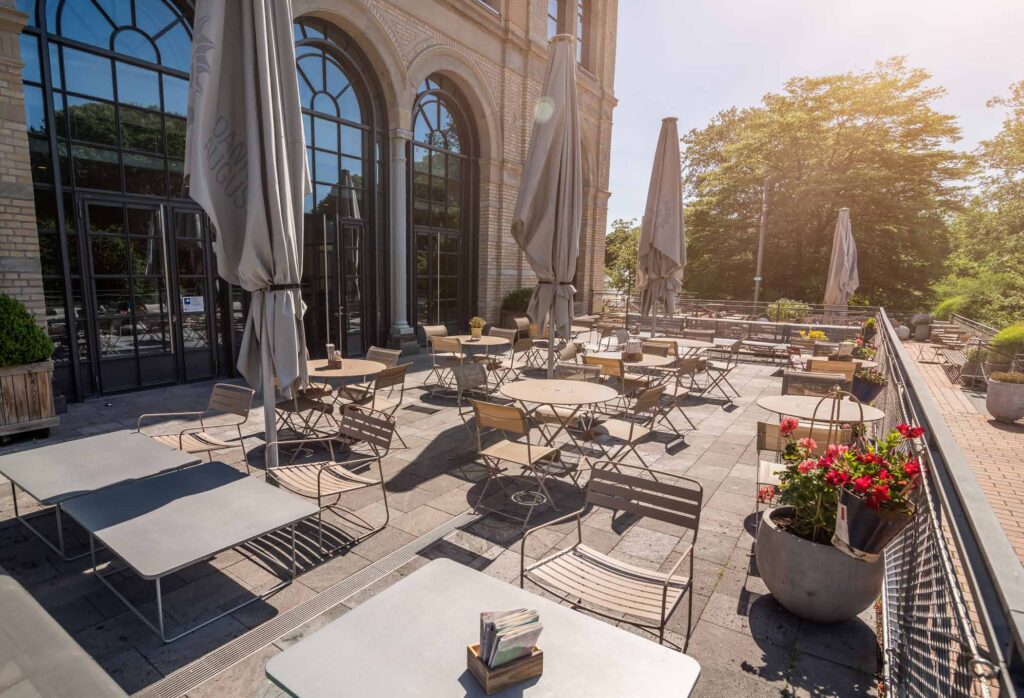 Sonnige Außenterrasse mit leeren Tischen, Stühlen, großen Topfpflanzen und geschlossenen Schirmen in der Nähe eines Gebäudes - perfekt für ein Catering in der Flora Köln.