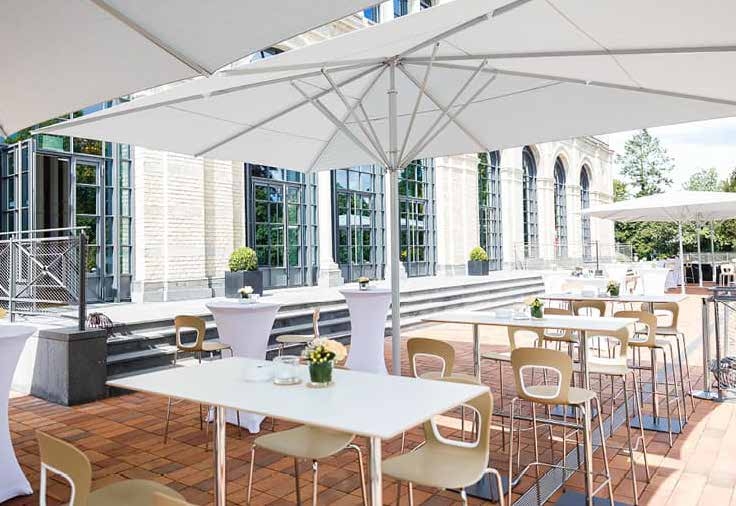 Eine Außenterrasse mit Tischen, Stühlen und großen Sonnenschirmen in der Nähe eines Gebäudes mit hohen Fenstern und Backsteintreppen - ideal, um das Catering in der Flora Köln zu genießen.