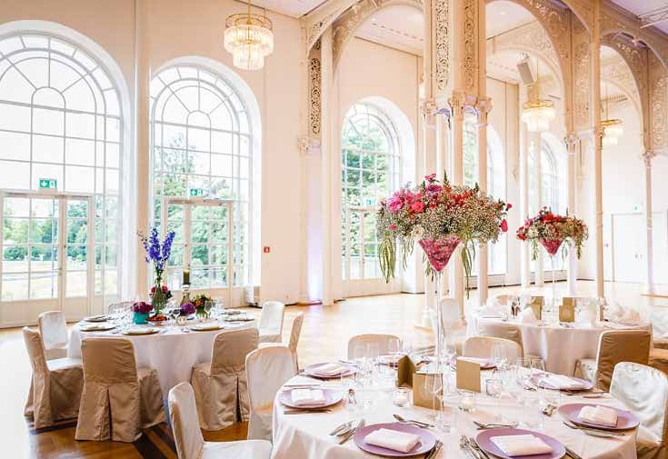 Eleganter Ballsaal mit hohen Bogenfenstern, runden Tischen und floralen Tafelaufsätzen für eine Veranstaltung oder Hochzeit - perfekt für alle, die Catering in der Flora Köln suchen.