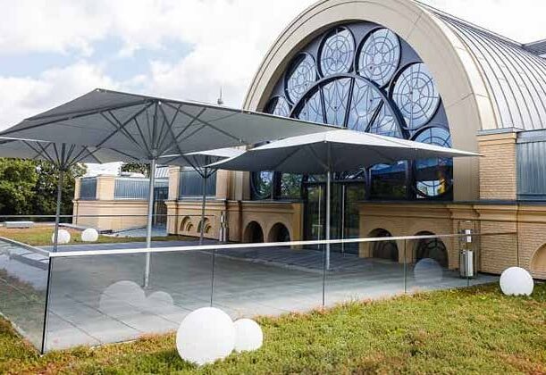 Modernes Gebäude mit großen Rundbogenfenstern, Terrassenschirmen und kugelförmigen Außenleuchten auf einer grünen Terrasse - perfekt für Veranstaltungen mit Catering in der Flora Köln.