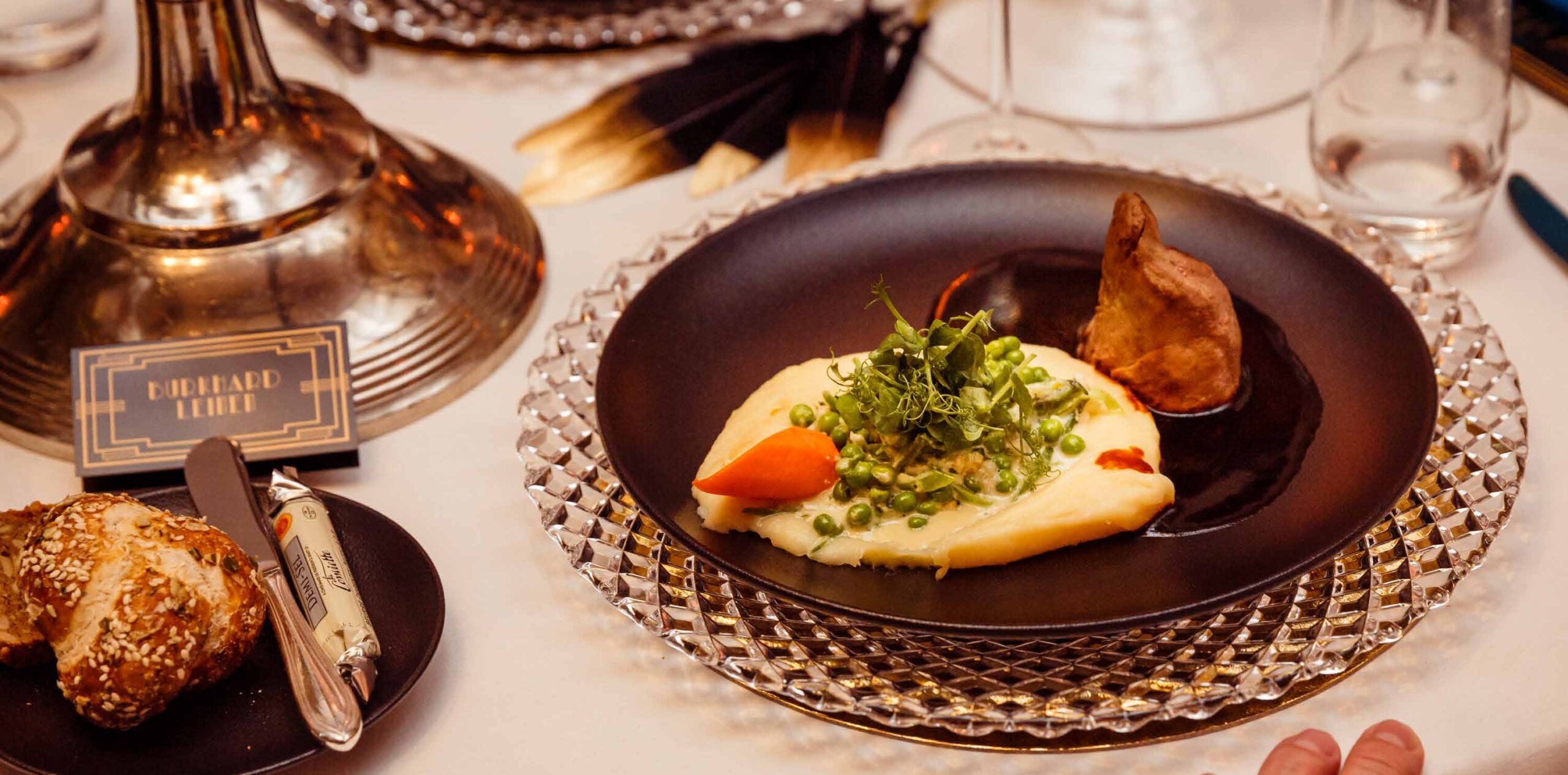 Elegantes Menü mit Kartoffelpüree, Gemüse und Fleisch auf einer Glasplatte mit Brötchen und Utensilien in der Nähe - perfekt für Eventcatering für besondere Anlässe.