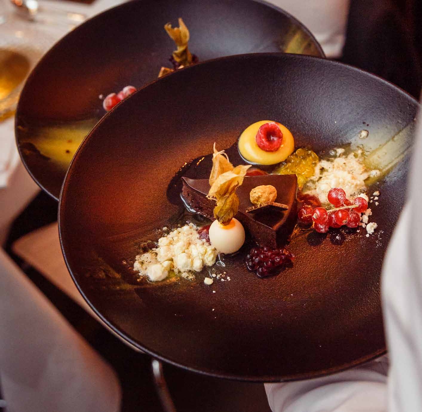 Elegantes Schokoladendessert mit Fruchtgarnituren und kunstvoller Präsentation auf einem schwarzen Teller - perfekt für Eventcatering für besondere Anlässe.