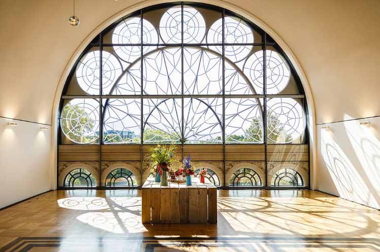 Ein großes Bogenfenster mit kreisrunden Mustern, einfallendes Sonnenlicht und ein Blumenarrangement auf einem Holztisch - so lässt sich das elegante Ambiente von Catering in der Flora Köln perfekt einfangen.