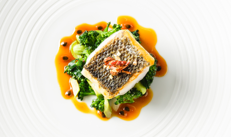Ein Fischfilet auf Grünzeug mit Orangensauce, garniert mit Kräutern, serviert auf einem weißen Teller - perfekt für professionelles Catering für Events in Köln.