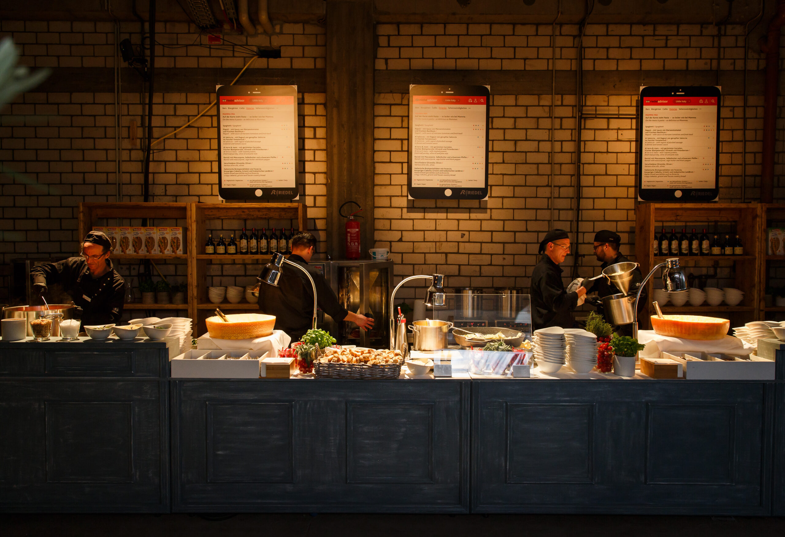 Drei Köche bereiten die Speisen hinter einer Theke mit Tellern und Käselaiben in einem warm beleuchteten Restaurant zu, ideal für Eventcatering für besondere Anlässe.