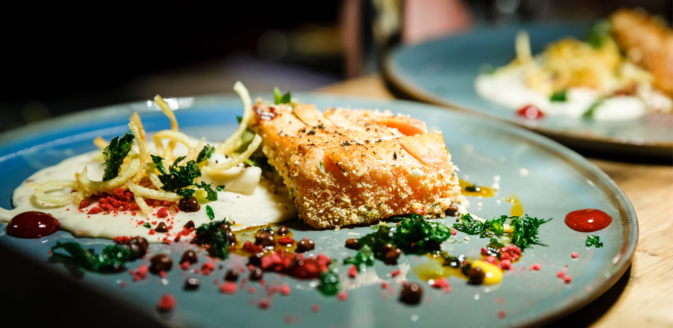 Ein Gourmet-Lachsfilet mit Sauce, knusprigen Zwiebeln und bunten Beilagen auf einem blauen Teller - perfekt für Eventcatering für besondere Anlässe.