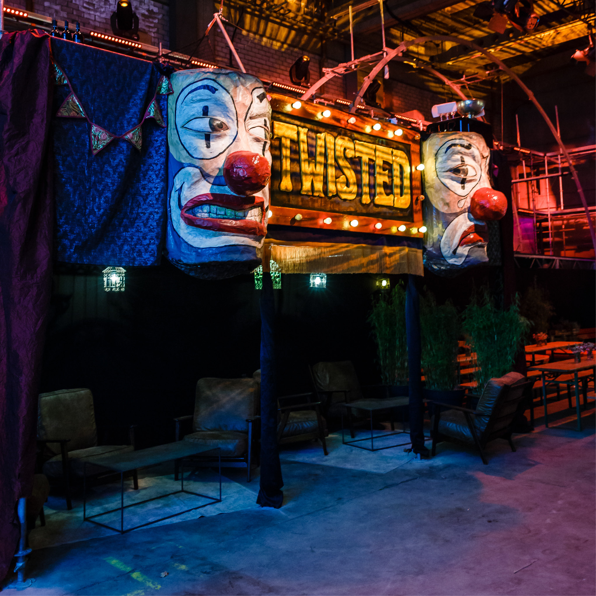 Eine zirkusähnliche Lounge mit Clownsgesichtern, einem TWISTED-Schild und bunten Lichtern in einer industriellen Umgebung - ideal für Eventcatering für besondere Anlässe.