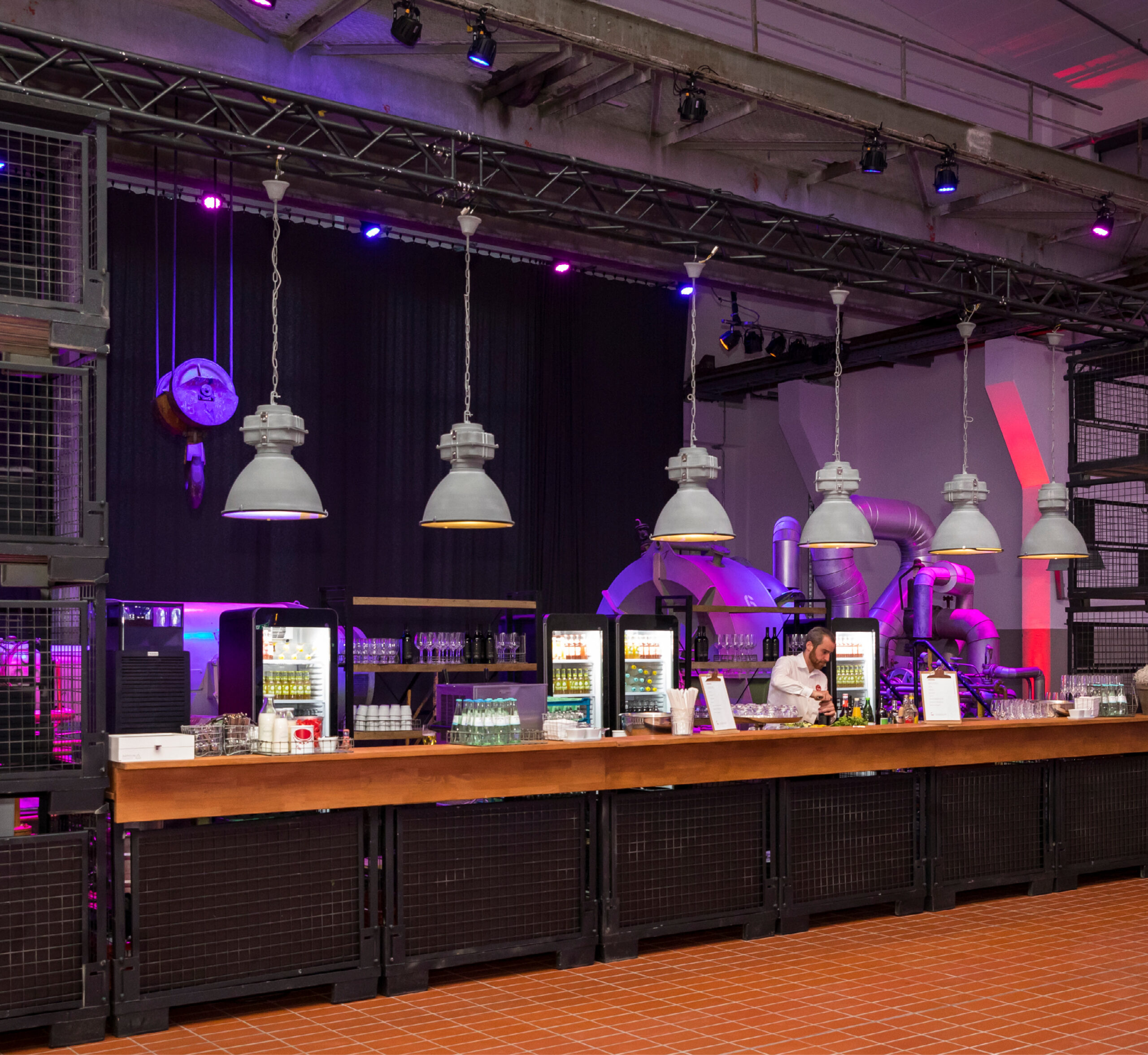 Bar im Industriestil mit Hängelampen, gefüllten Getränkekühlschränken und einem Barkeeper hinter einem Holztresen - ideal für Eventcatering für besondere Anlässe.