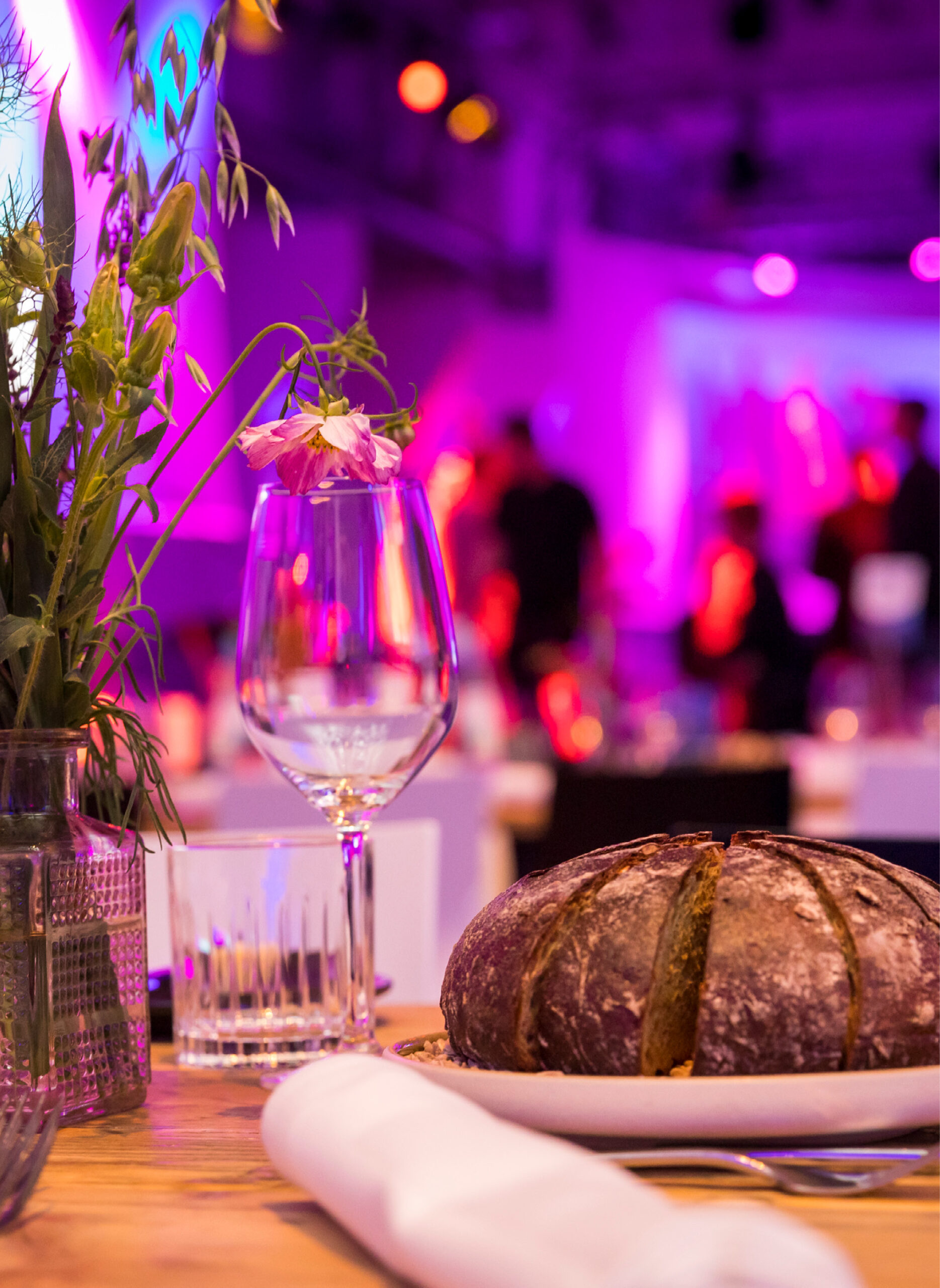 Ein rustikaler runder Brotlaib auf einem Teller mit einem Weinglas und Blumen auf einem Tisch bei einem lebhaften Eventcatering für besondere Anlässe.