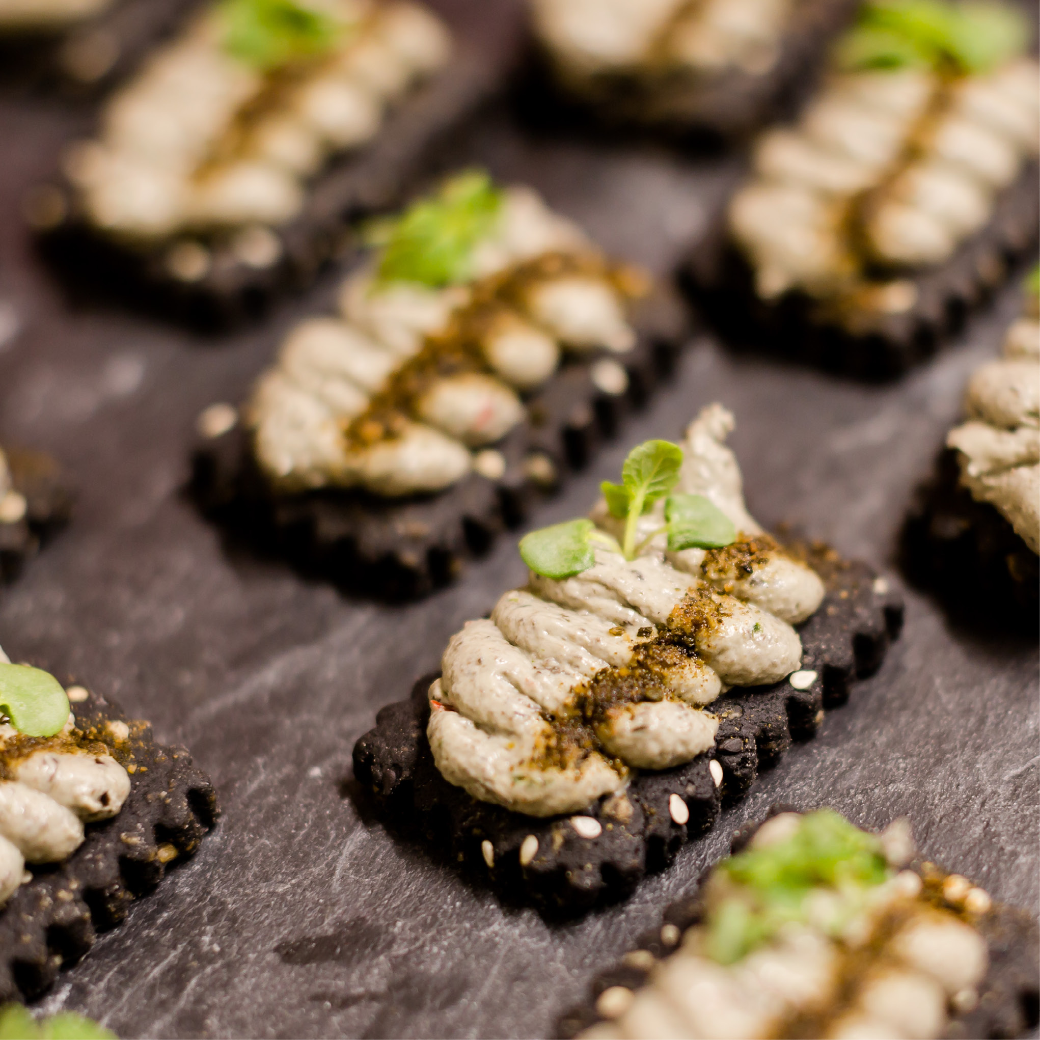 Rechteckige schwarze Cracker, belegt mit einem cremigen Aufstrich, Kräutern und Gewürzen, angeordnet auf einer dunklen Oberfläche - ideal für Eventcatering für besondere Anlässe.
