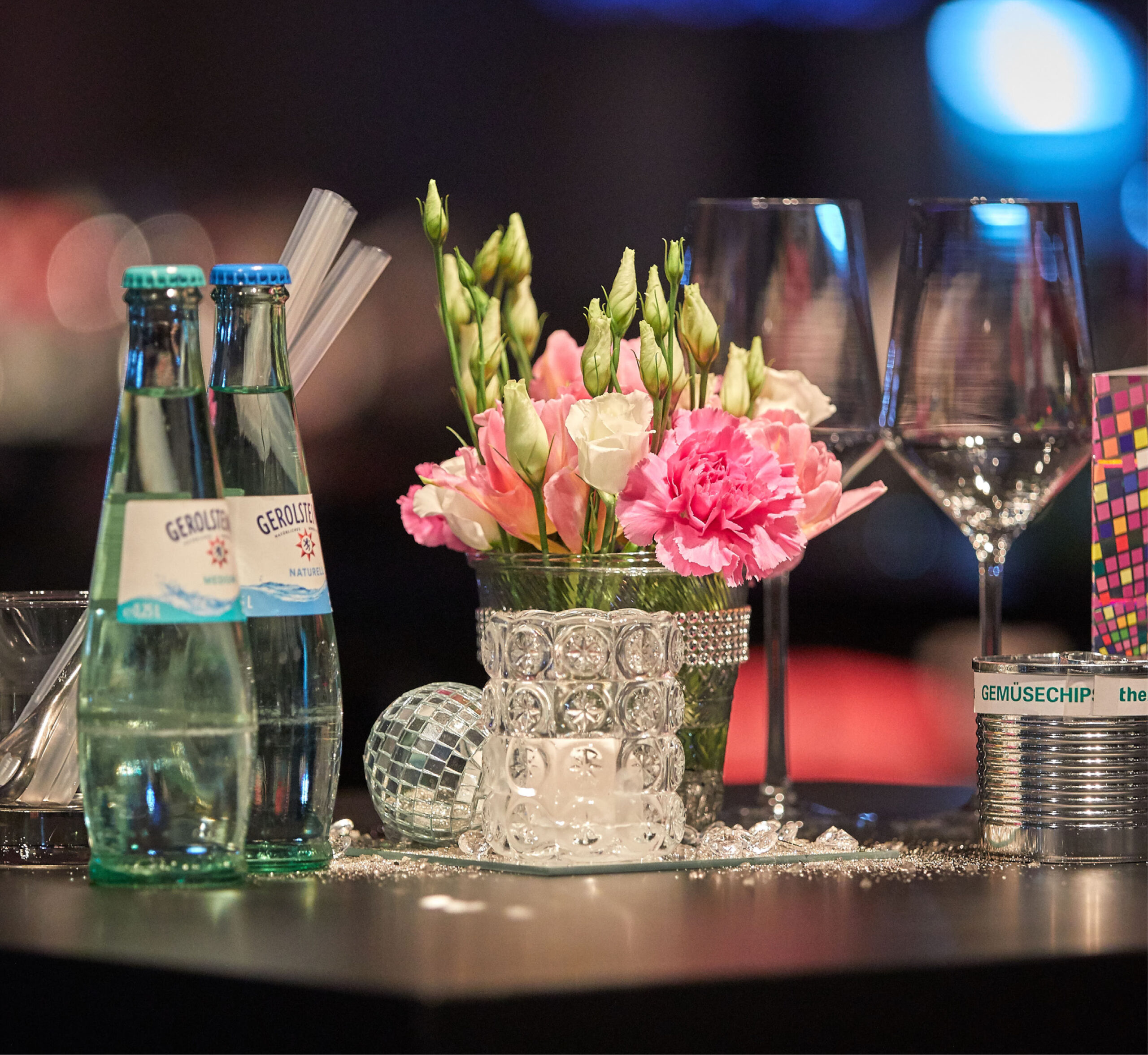 Glasflaschen, rosafarbene Blumen in einer Vase, Gläser und Strohhalme auf einem Tisch mit verstreuten Dekorationen - perfekt für Eventcatering für besondere Anlässe.