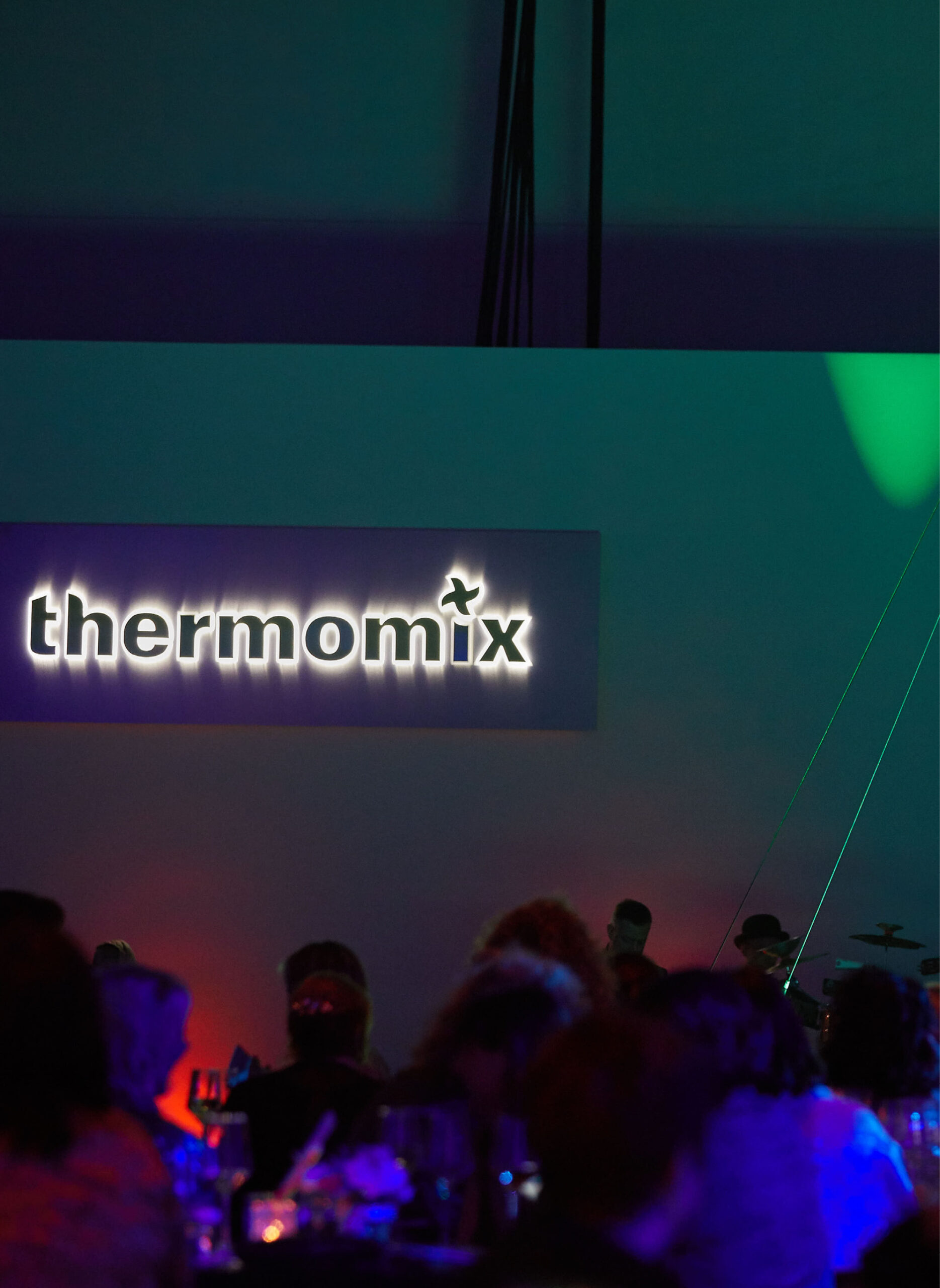 Schummerig beleuchtete Veranstaltung mit sitzenden Personen, die auf ein beleuchtetes Thermomix-Schild an der Wand blicken - perfekt für Eventcatering für besondere Anlässe.