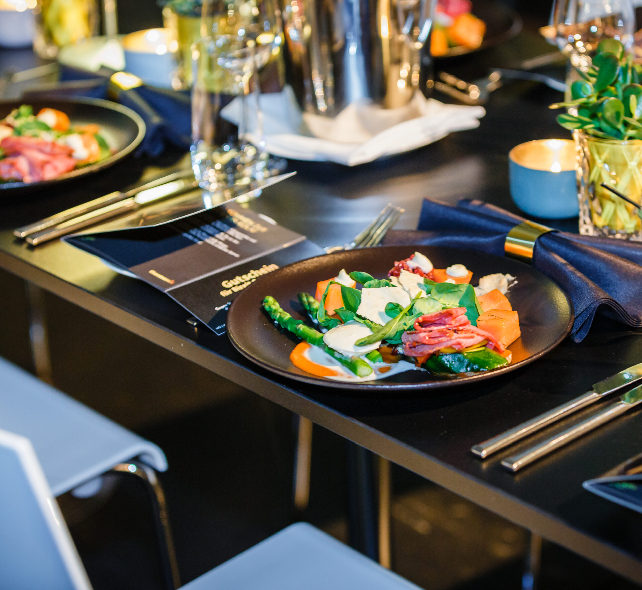 Ein Gourmet-Menü auf einem schwarz gedeckten Tisch mit Besteck, Gläsern und einer marineblauen Serviette - perfekt für Eventcatering für besondere Anlässe.