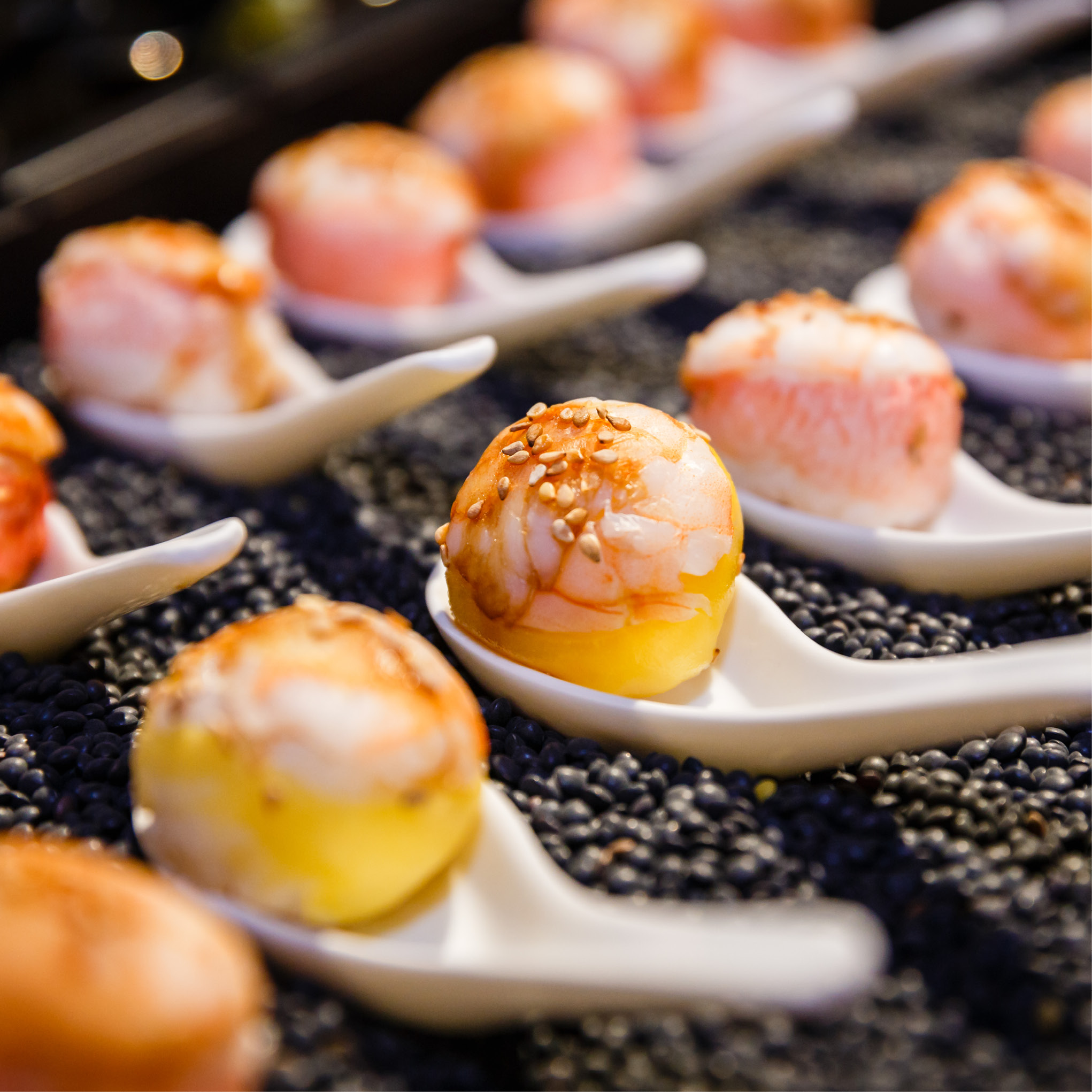Runde Sushi-Stücke mit Garnelen und Sauce, serviert auf weißen Keramiklöffeln und angerichtet auf einem Bett aus schwarzen Bohnen - perfekt für Eventcatering für besondere Anlässe.