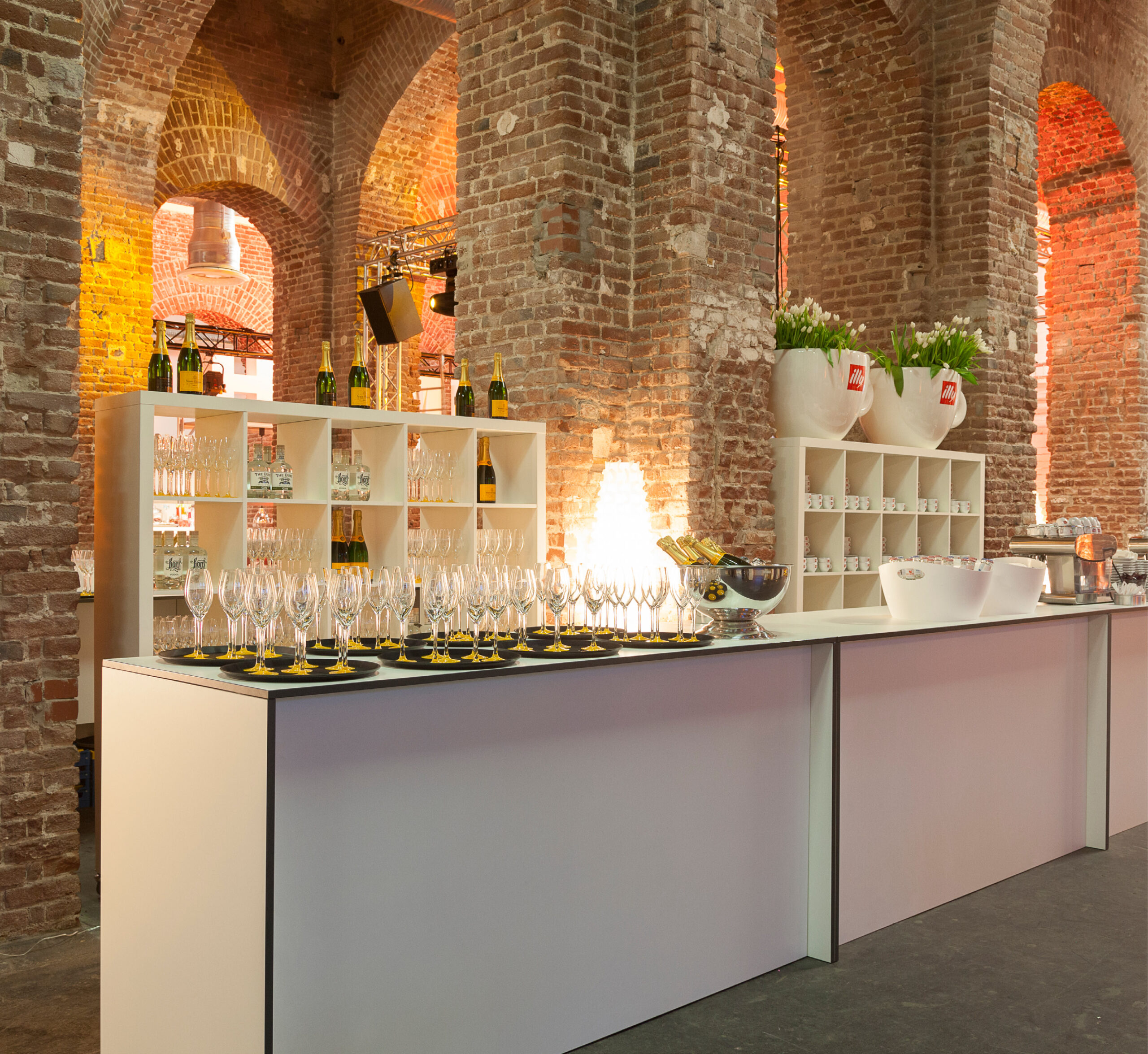 Modernes Bar-Setup mit vielen Weingläsern auf einem Tresen, in einem gemauerten Gewölbe mit Regalen und Dekoration-perfekt für Eventcatering für besondere Anlässe.