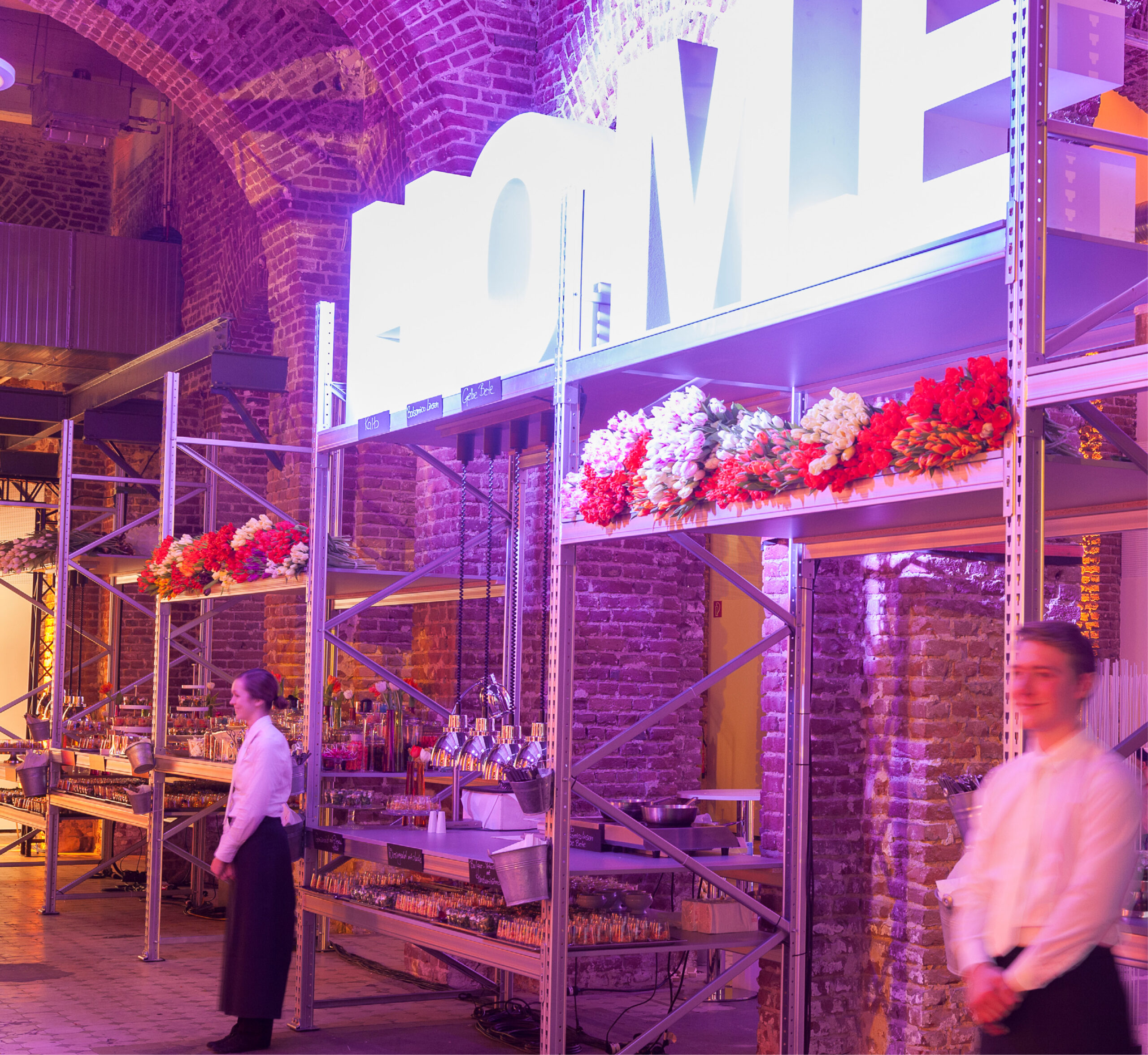 Das Catering-Personal steht an Tischen mit Blumen und großen Leuchtbuchstaben in einer gewölbten Backsteinhalle - perfektes Eventcatering für besondere Anlässe.