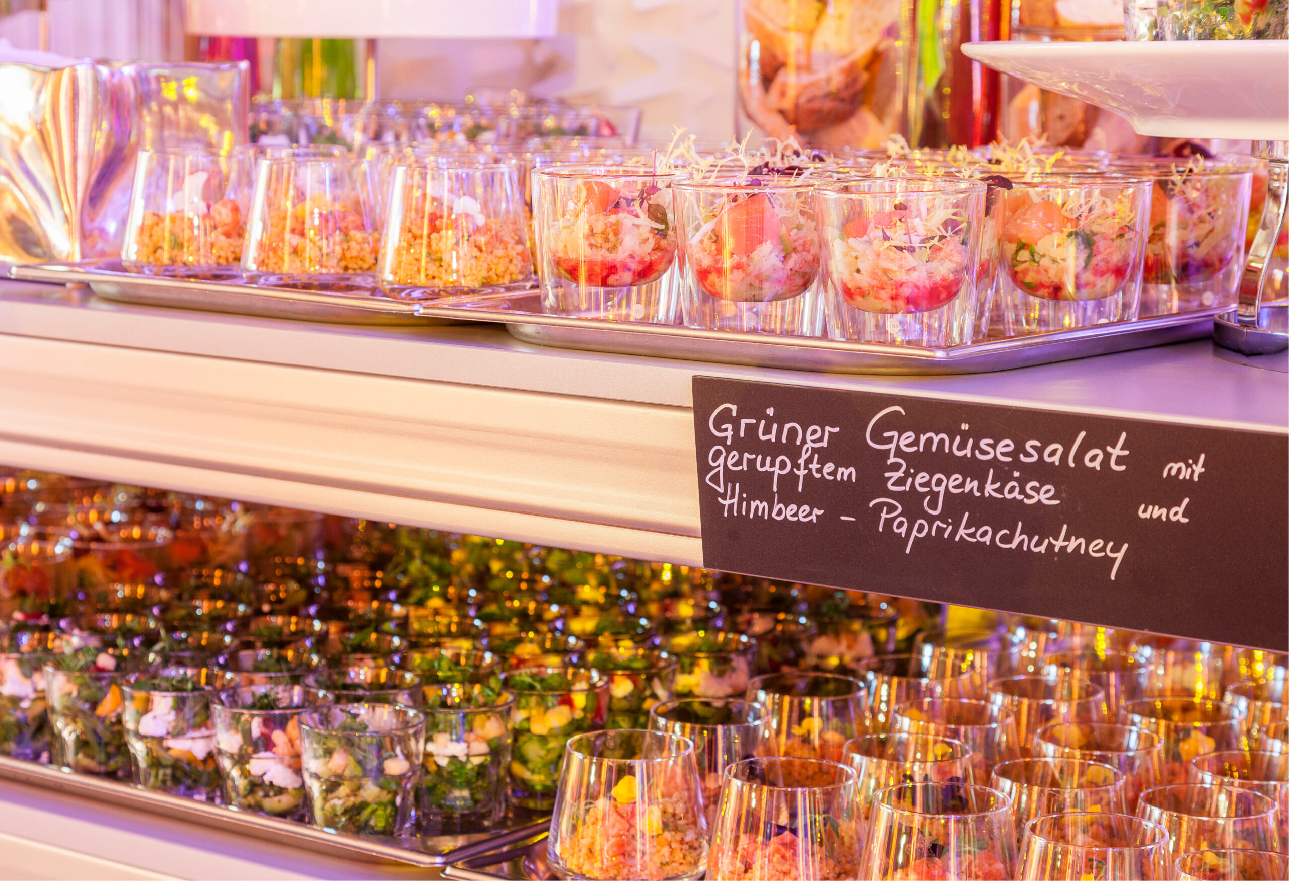 Gemischte Salate in Glasbechern mit einem Kreideschild, das den grünen Gemüsesalat beschreibt, perfekt für Eventcatering für besondere Anlässe.