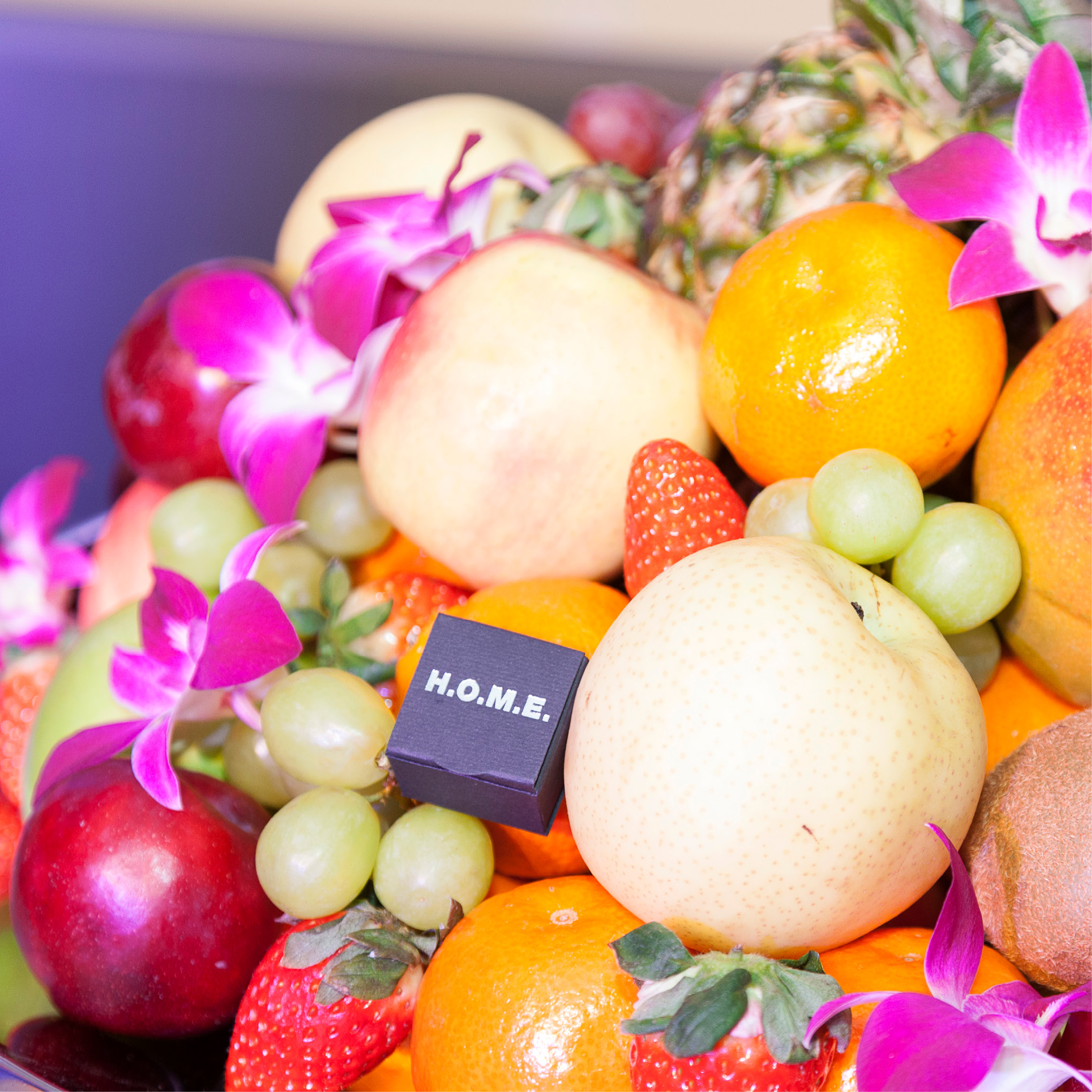 Ein farbenfrohes Obstarrangement mit lila Blumen und einem kleinen schwarzen Block mit der Aufschrift H.O.M.E. - perfekt für Eventcatering für besondere Anlässe.