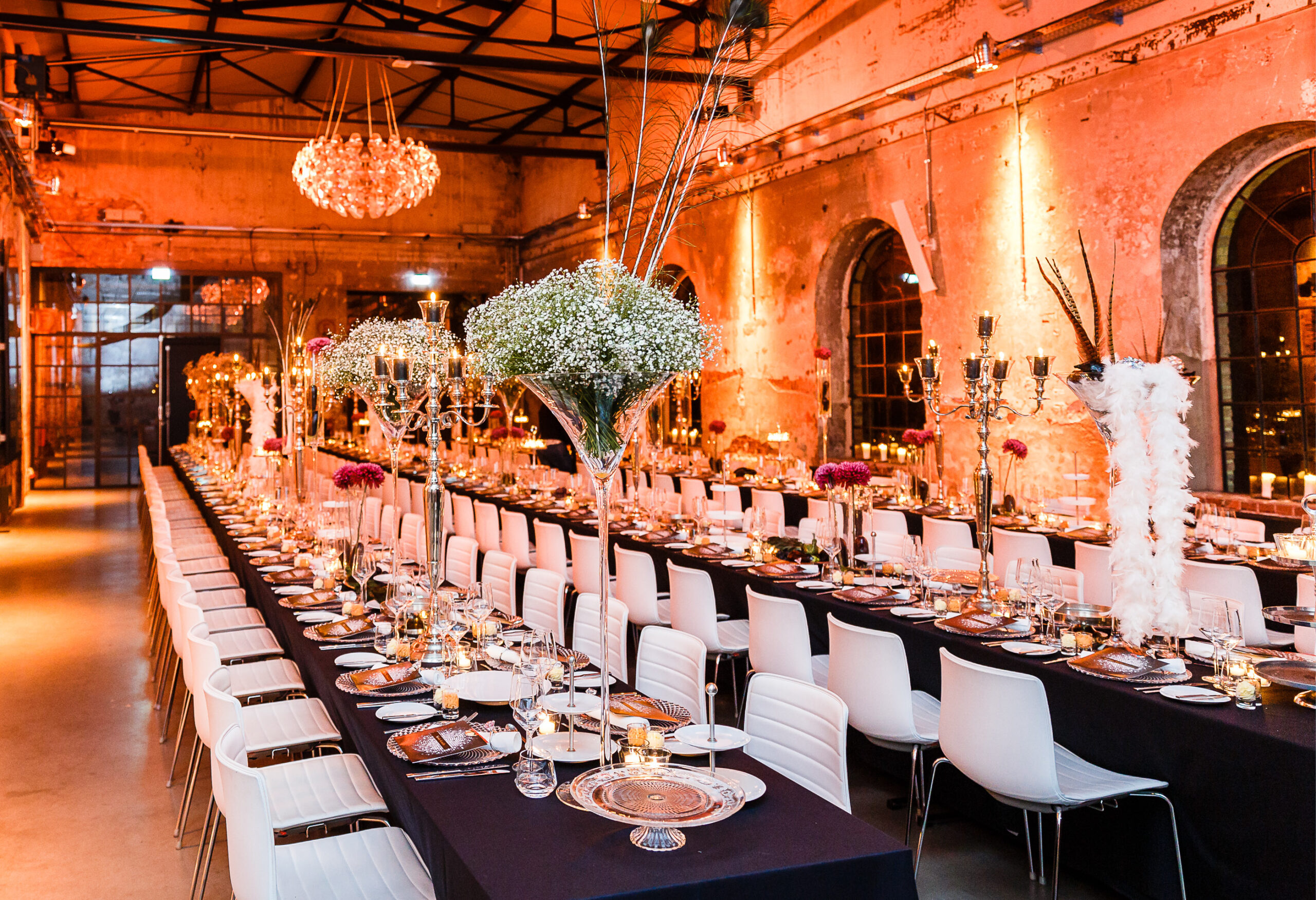 Eleganter Bankettsaal mit langen Tischen, weißen Stühlen, Kerzen und floralen Tafelaufsätzen, warm beleuchtet - perfekt für Fine Dining Catering in Köln.
