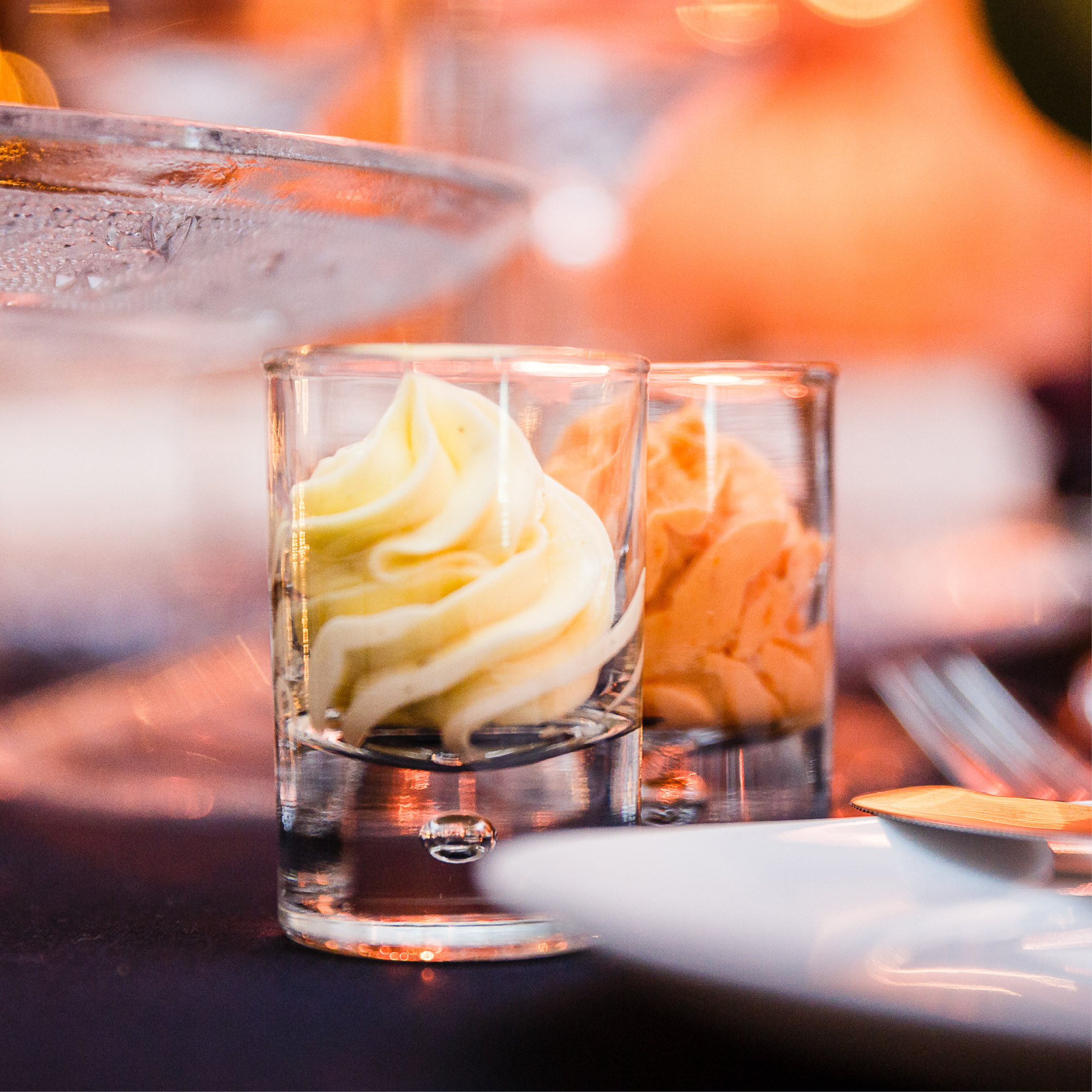 Zwei kleine Gläser, gefüllt mit Sahne- oder Mousse-Spritzern, stehen auf einem Tisch und zeigen die Eleganz von Fine Dining Catering in Köln vor einem sanft verschwommenen Hintergrund.