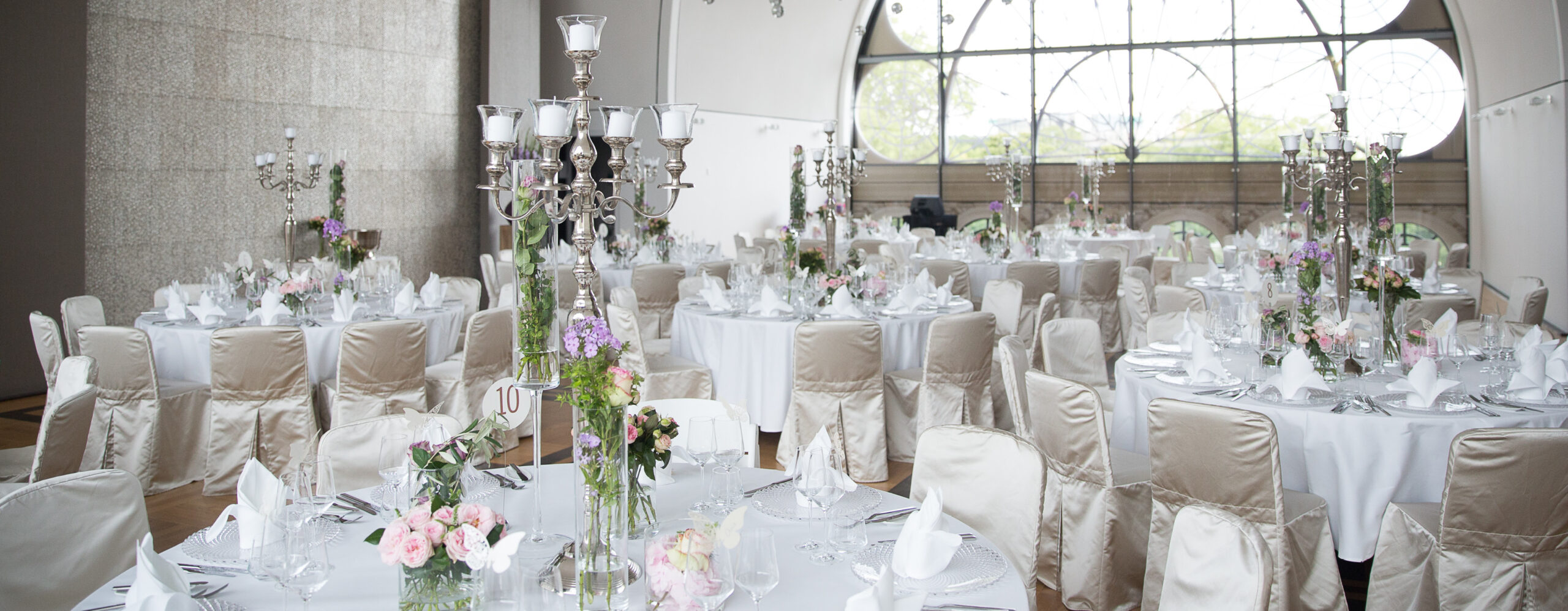 Eleganter Bankettsaal mit runden Tischen, weißem Leinen, Blumenschmuck und Kandelabern für eine Veranstaltung. Ideal für Catering für Hochzeiten und Feiern, verspricht dieser Raum eine unvergessliche Feier in einer raffinierten Atmosphäre.