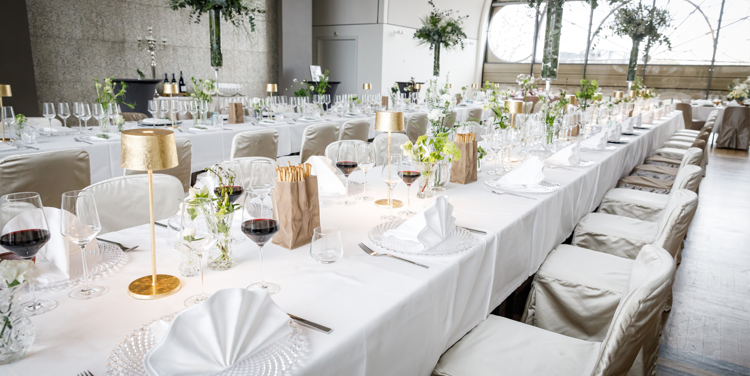 Elegante Banketttische mit weißer Bettwäsche, goldenen Lampen, Gläsern und floralen Tafelaufsätzen in einem hellen Raum - perfekt für Catering für Hochzeiten und Feiern.