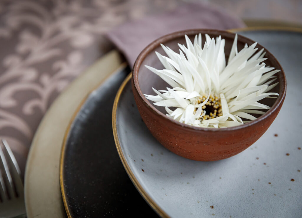 Eine braune Schale mit einer weißen Blume darin, auf gestapelten Tellern mit einem gemusterten Tuch daneben - perfekt für Catering für Hochzeiten und Feiern.