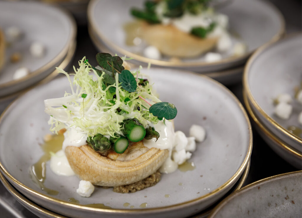Eine plattierte Vorspeise mit Blätterteig, frischem Grün, Spargel, Käse und cremiger Sauce auf einer weißen Platte - perfekt für Catering für Hochzeiten und Feiern.