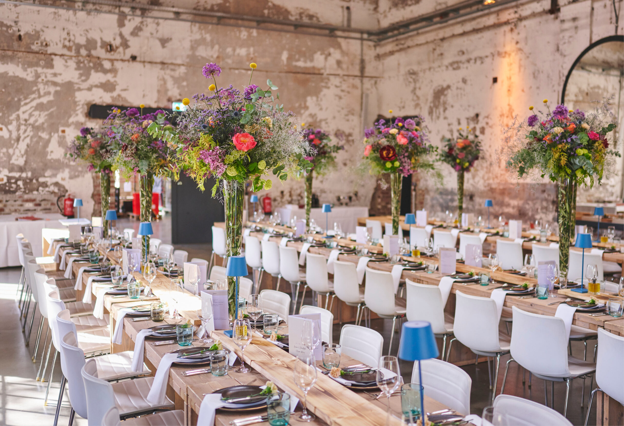 Lange Banketttische mit weißen Stühlen und hohe, farbenfrohe Blumenkränze bilden den Rahmen für Catering für Hochzeiten und Feiern in einer rustikalen Industriehalle.