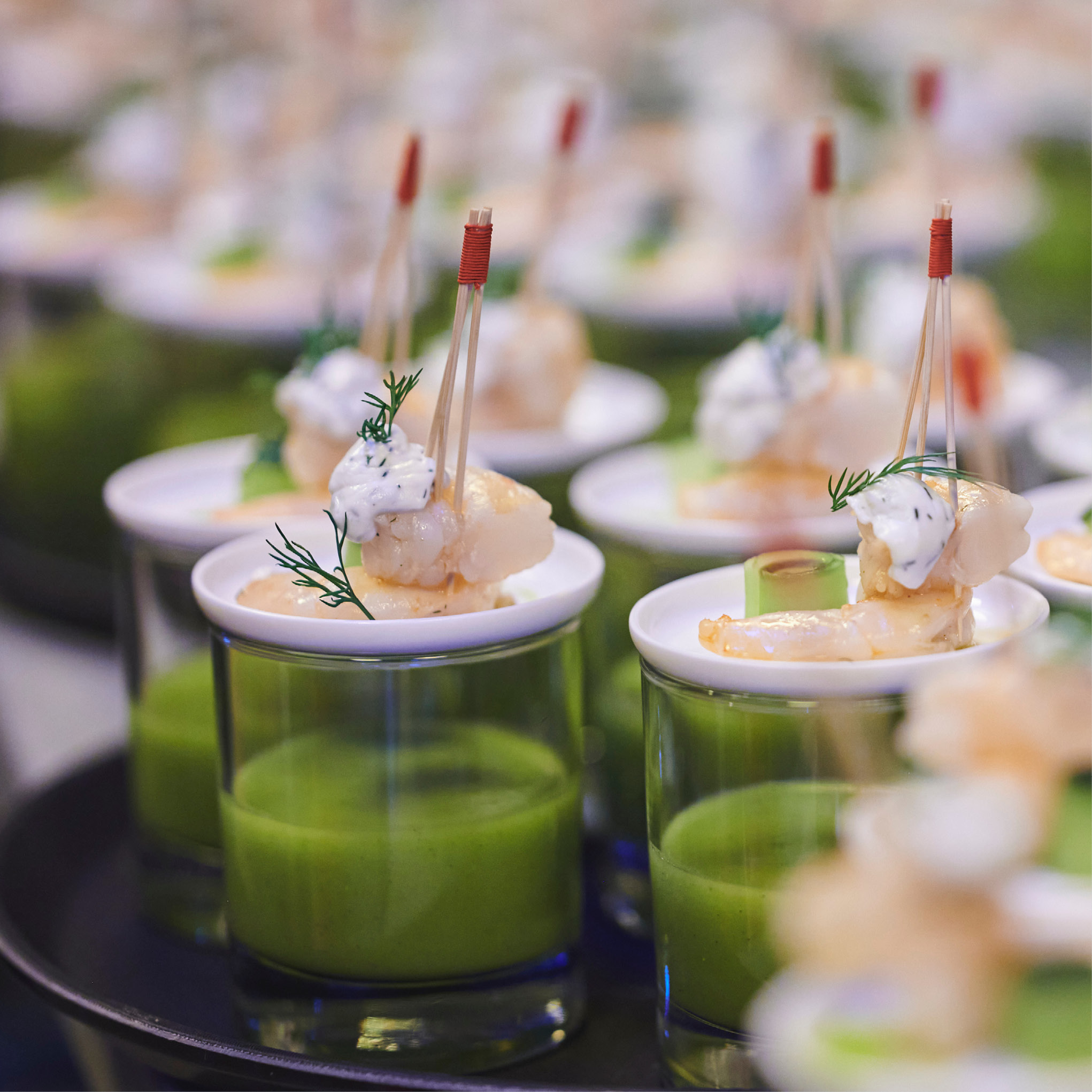 Kleine Gläser mit grüner Suppe, belegt mit aufgespießten Garnelen und Kräutern, serviert als elegante Vorspeise - perfekt für Catering für Hochzeiten und Feiern.