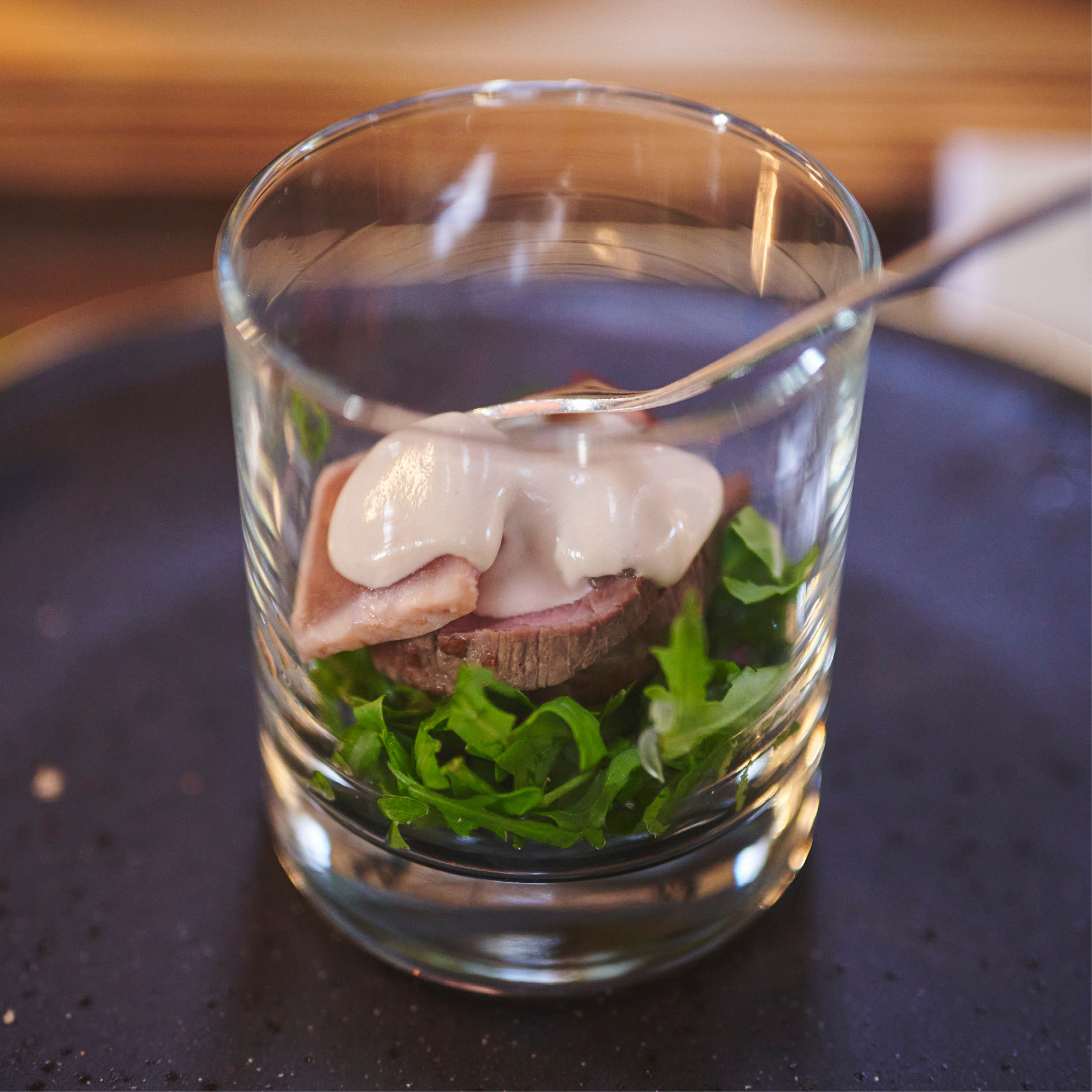 Ein Glas mit Grünzeug, Aufschnitt und einer cremigen weißen Soße obenauf, schön serviert auf einem dunklen Teller - ideal für Catering für Hochzeiten und Feiern.