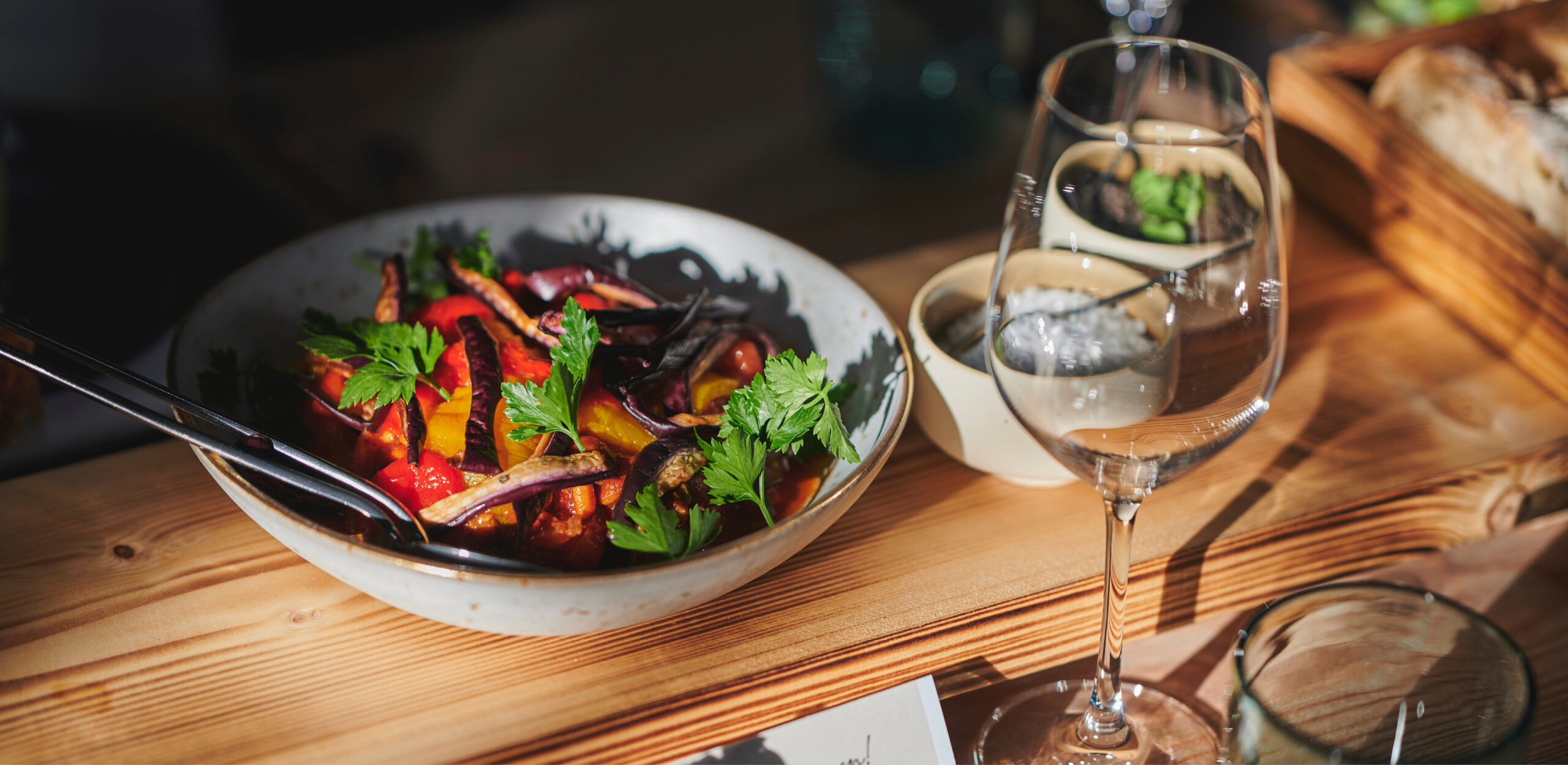 Auf einem Holztisch steht eine Schale mit buntem, gebratenem Gemüse und Kräutern, daneben leere Weingläser - perfekt für das Catering für Hochzeiten und Feiern.