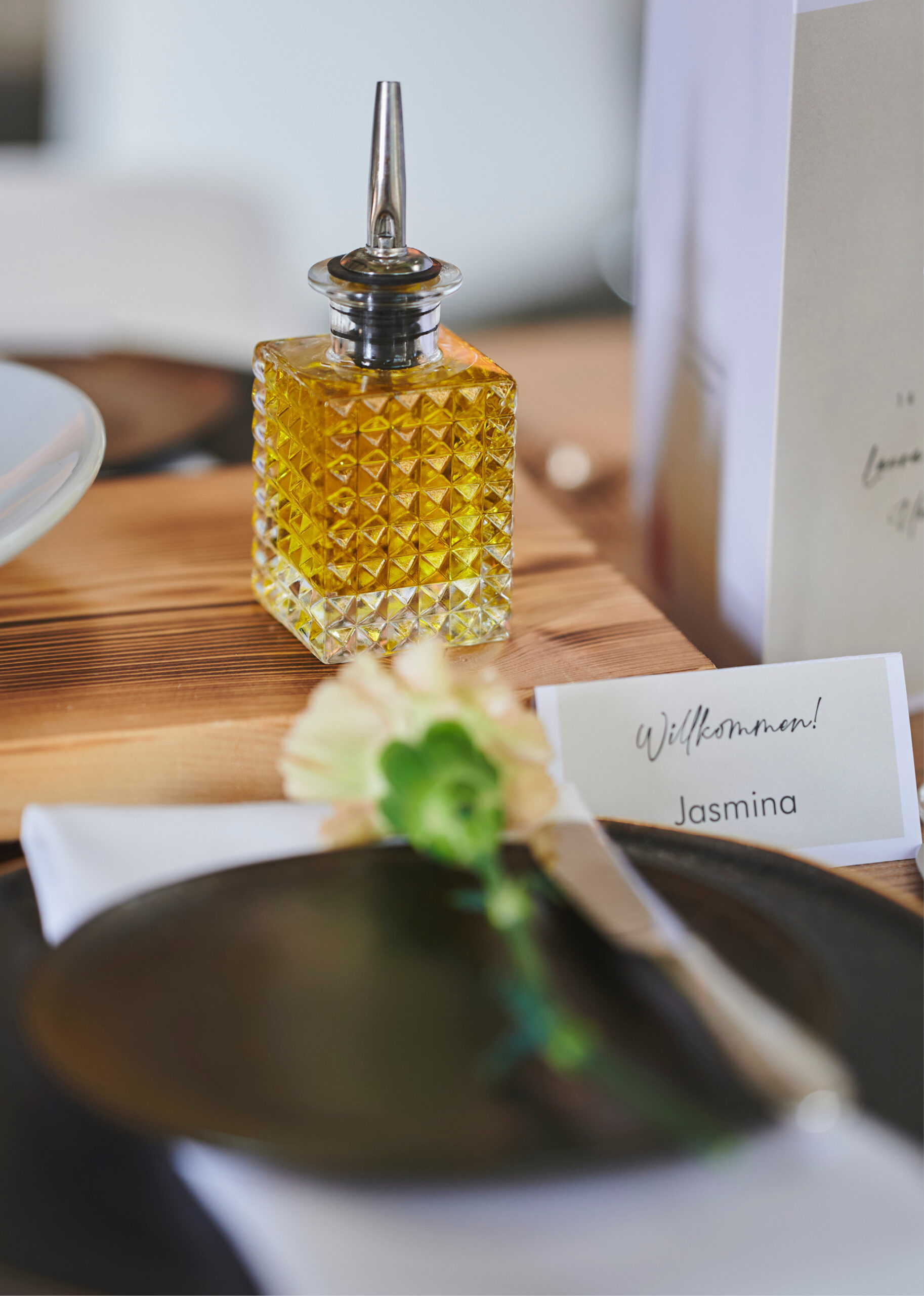 Glasflasche mit gelber Flüssigkeit neben einem Namensschild mit der Aufschrift Willkommen! Jasmina an einem gedeckten Esstisch - perfekt für Catering für Hochzeiten und Feiern.