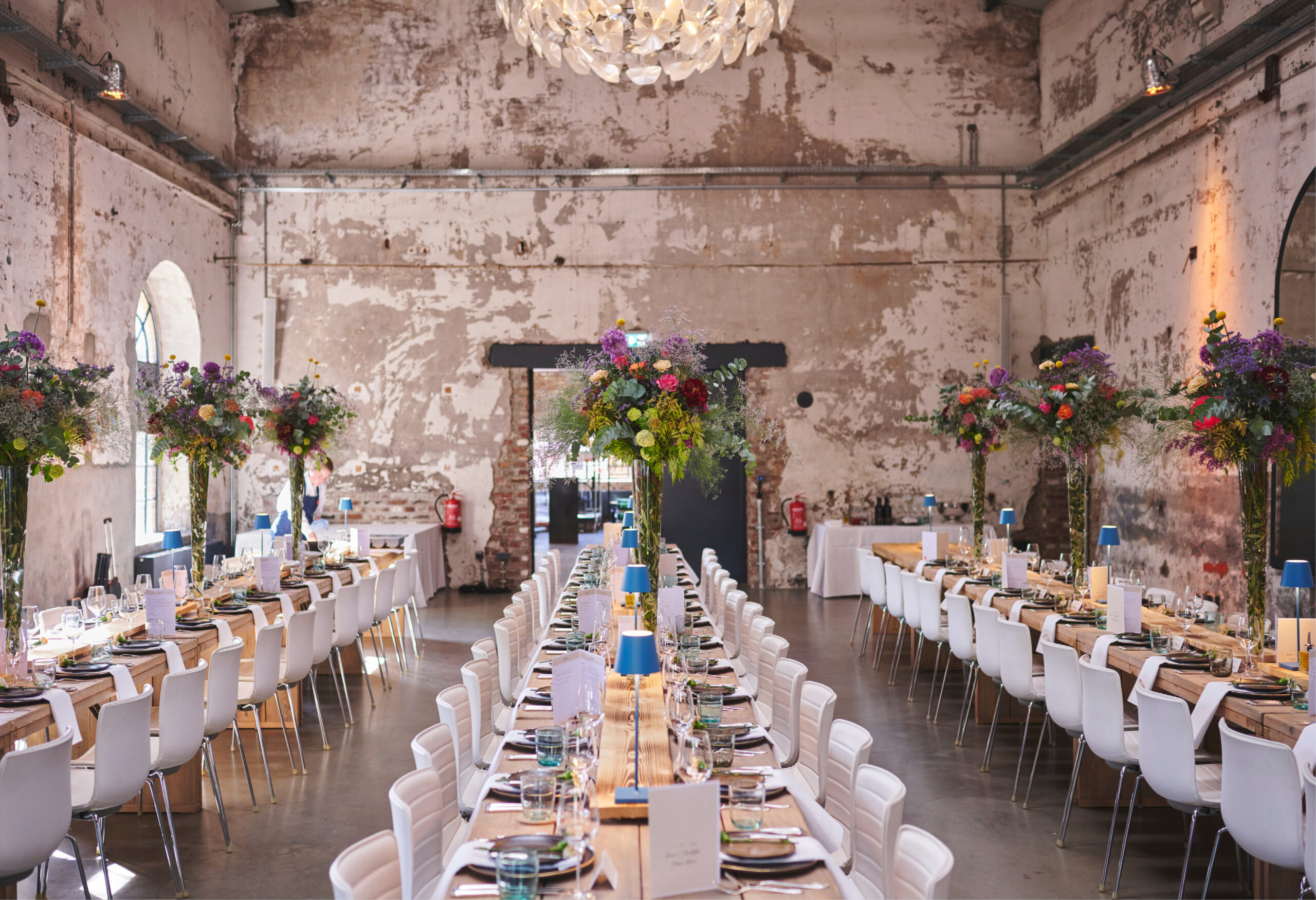 Eine lange Banketttafel mit eleganten Gedecken und hohen Blumensträußen in einem Lokal im Industriestil, perfekt für Catering für Hochzeiten und Feiern.