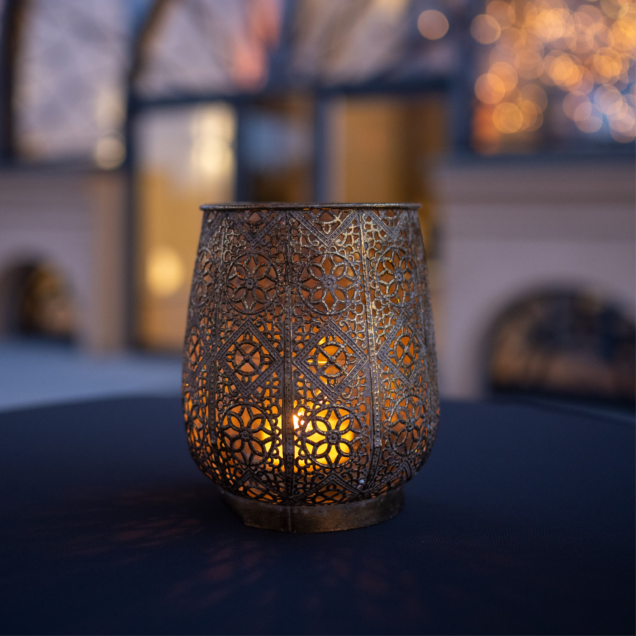 Verzierte Metalllaterne mit einer brennenden Kerze im Inneren, die auf einem dunklen Tisch warm leuchtet - perfekt für ein stimmungsvolles Catering für Hochzeiten und Feiern, mit einem schön verschwommenen Hintergrund.