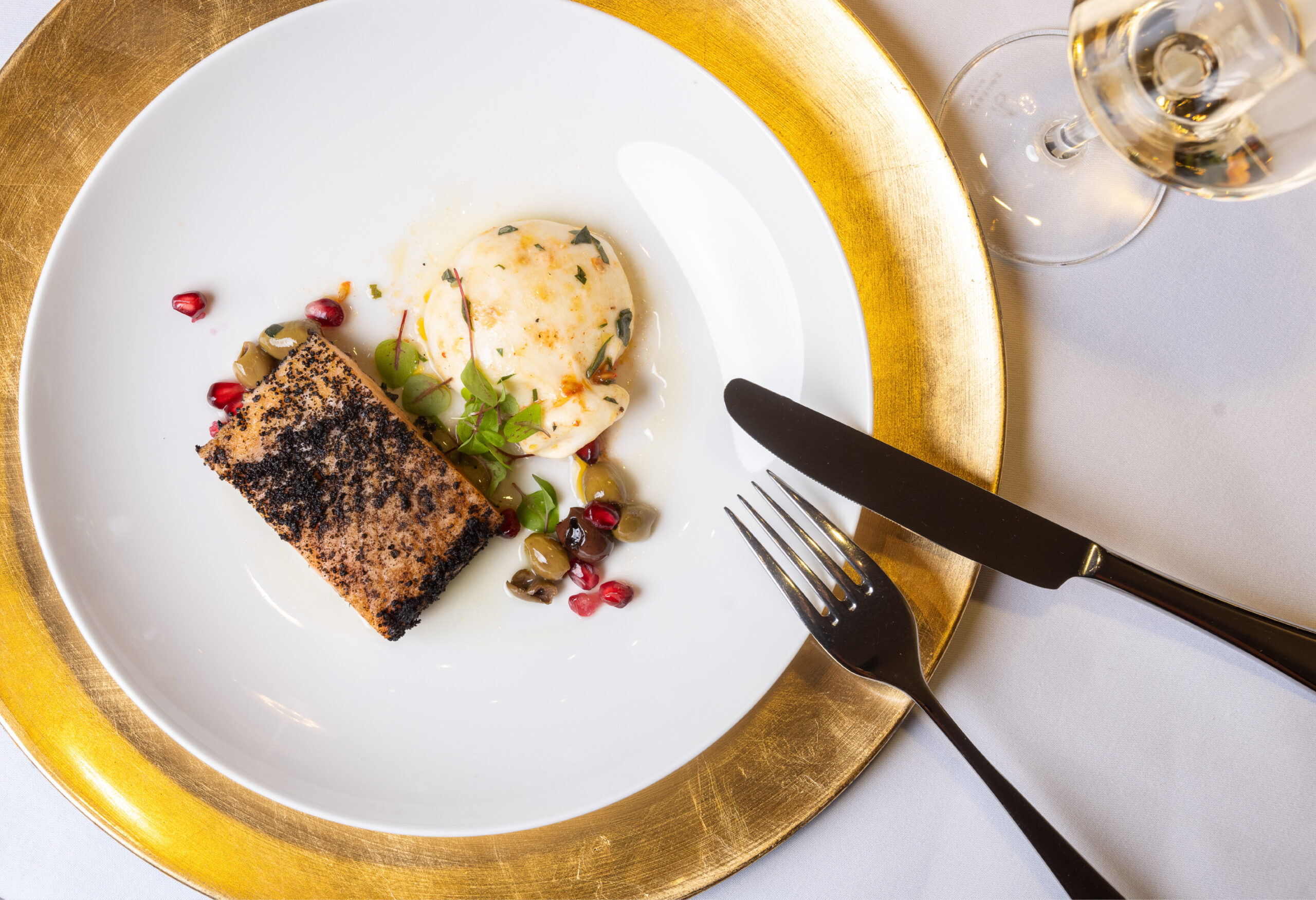 Ein plattiertes Gourmet-Menü mit Fisch, Sauce, Oliven, Granatapfelkernen und einem Glas Weißwein - perfekt für das Catering für Hochzeiten und Feiern.