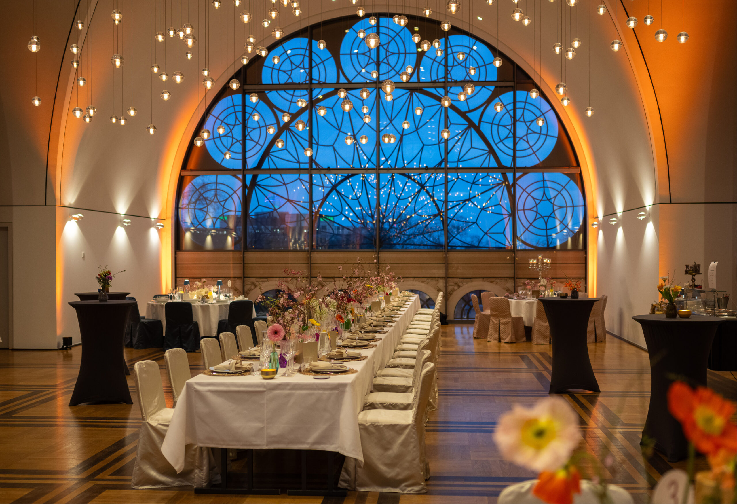 Eleganter Bankettsaal mit langen Tischen, floralen Tafelaufsätzen und einem großen Bogenfenster mit dekorativem Glas - perfekt für Catering für Hochzeiten und Feiern.
