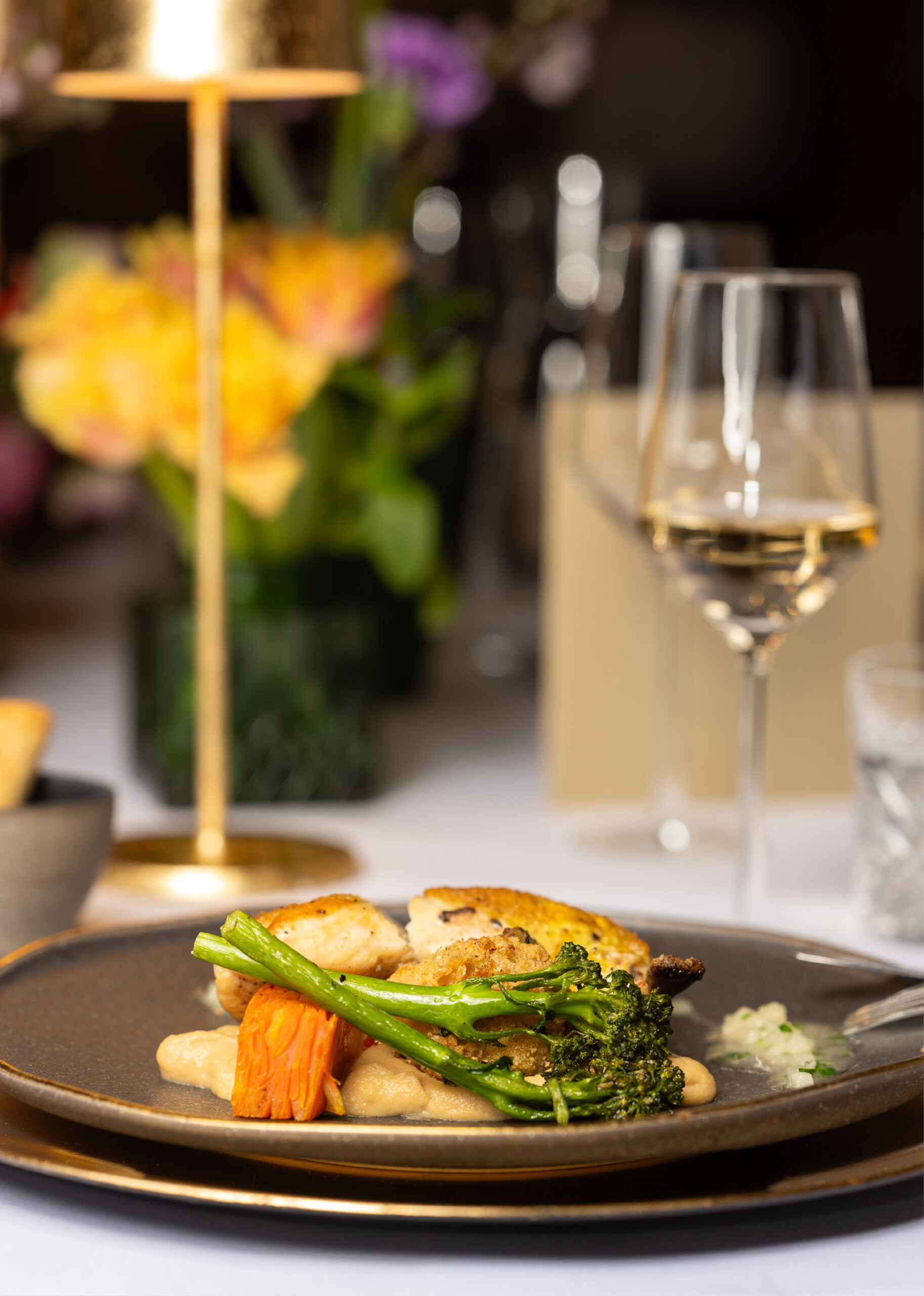Ein plattiertes Gourmet-Menü mit Gemüse und Huhn, perfekt für Catering für Hochzeiten und Feiern, wird neben einem Glas Weißwein und einer goldenen Lampe auf einem stilvollen Tisch serviert.