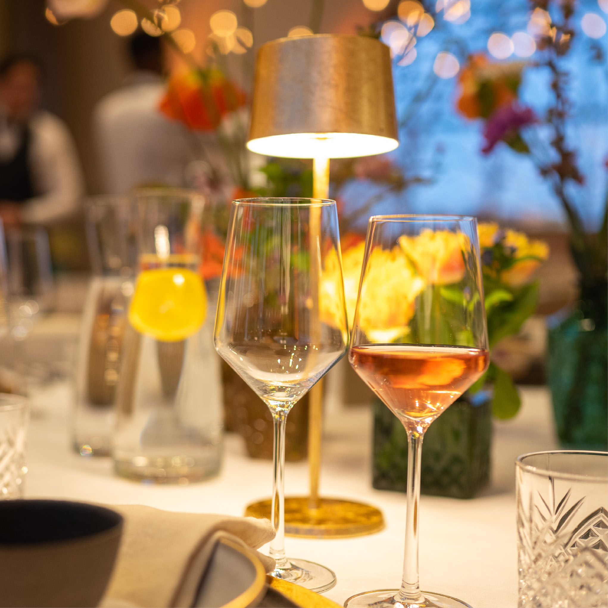 Zwei Weingläser auf einem dekorierten Tisch mit Blumen und einer warmen Lampe in einem gemütlichen, festlichen Rahmen - ideal für Catering für Hochzeiten und Feiern.