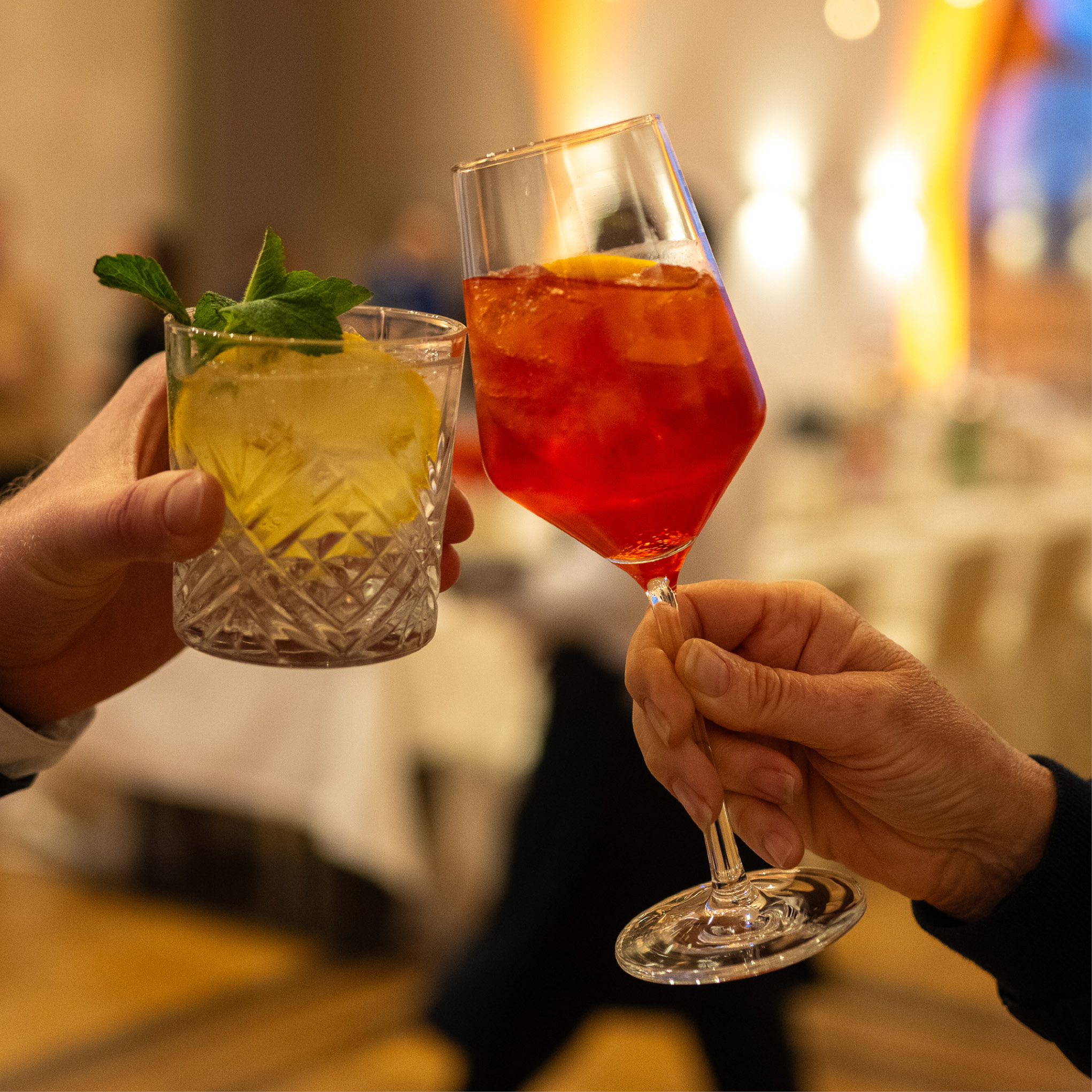 Zwei Personen stoßen mit Gläsern an - eine mit einem gelben Cocktail, die andere mit einem roten Getränk - und genießen die lebhafte Atmosphäre, die perfekt für Catering für Hochzeiten und Feiern ist.