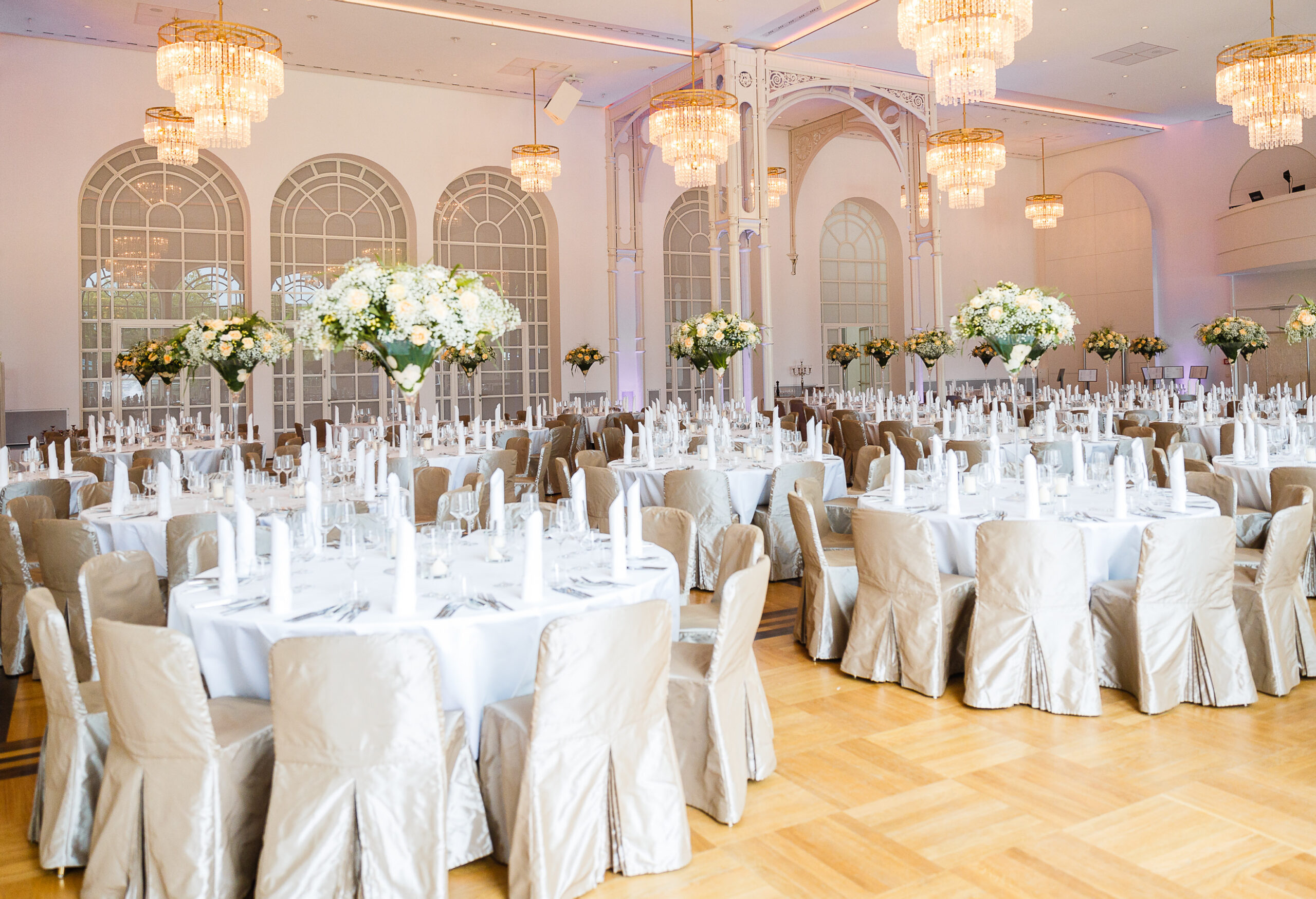Eleganter Festsaal mit runden Tischen, silbernen Stühlen, weißen Blumen und Kristalllüstern - ideal für Catering für Hochzeiten und Feiern.