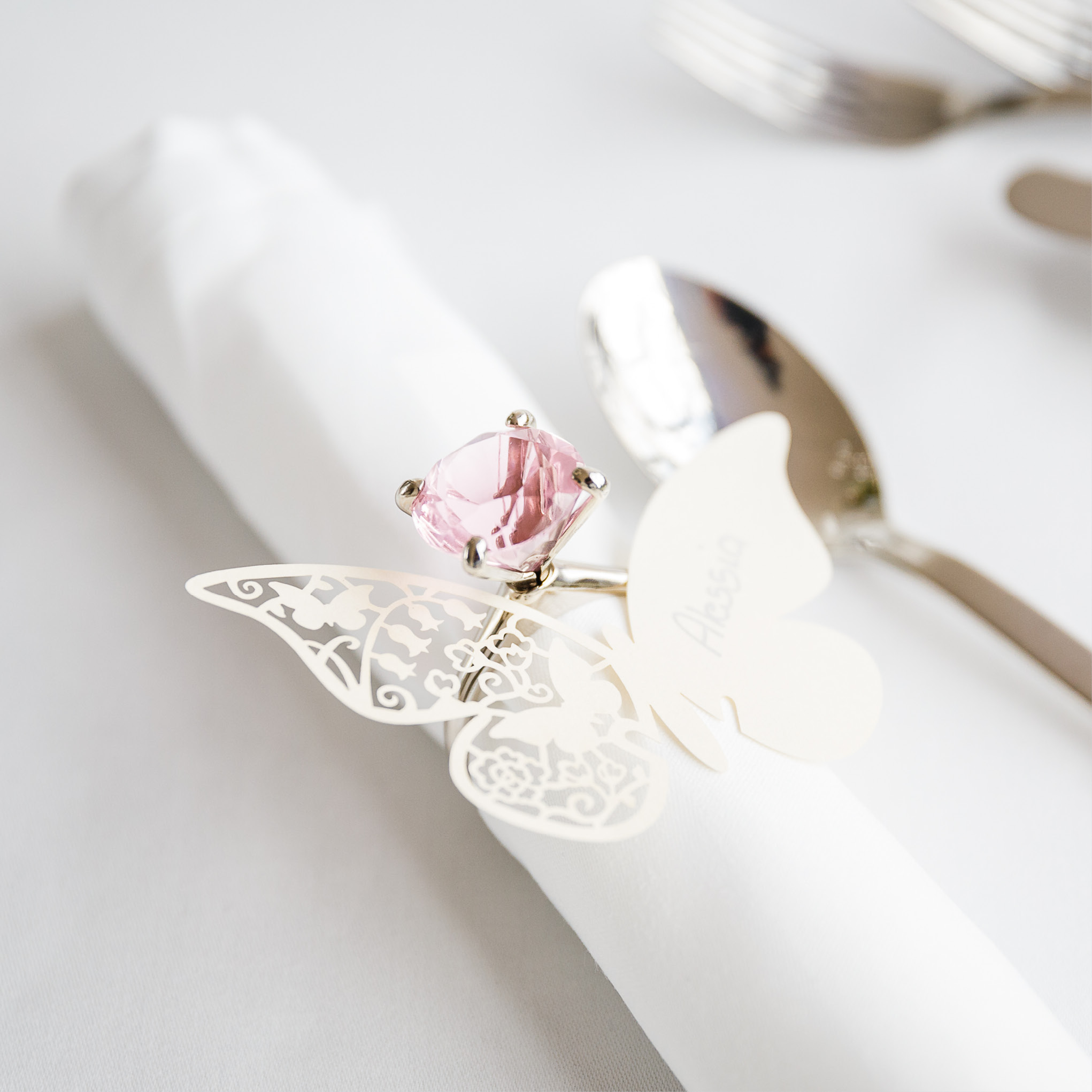 Weiße Serviette mit rosafarbenem Edelstein-Serviettenring und Namenskärtchen in Schmetterlingsform, daneben ein Löffel auf einem weißen Tisch - perfekt für elegantes Catering für Hochzeiten und Feiern.