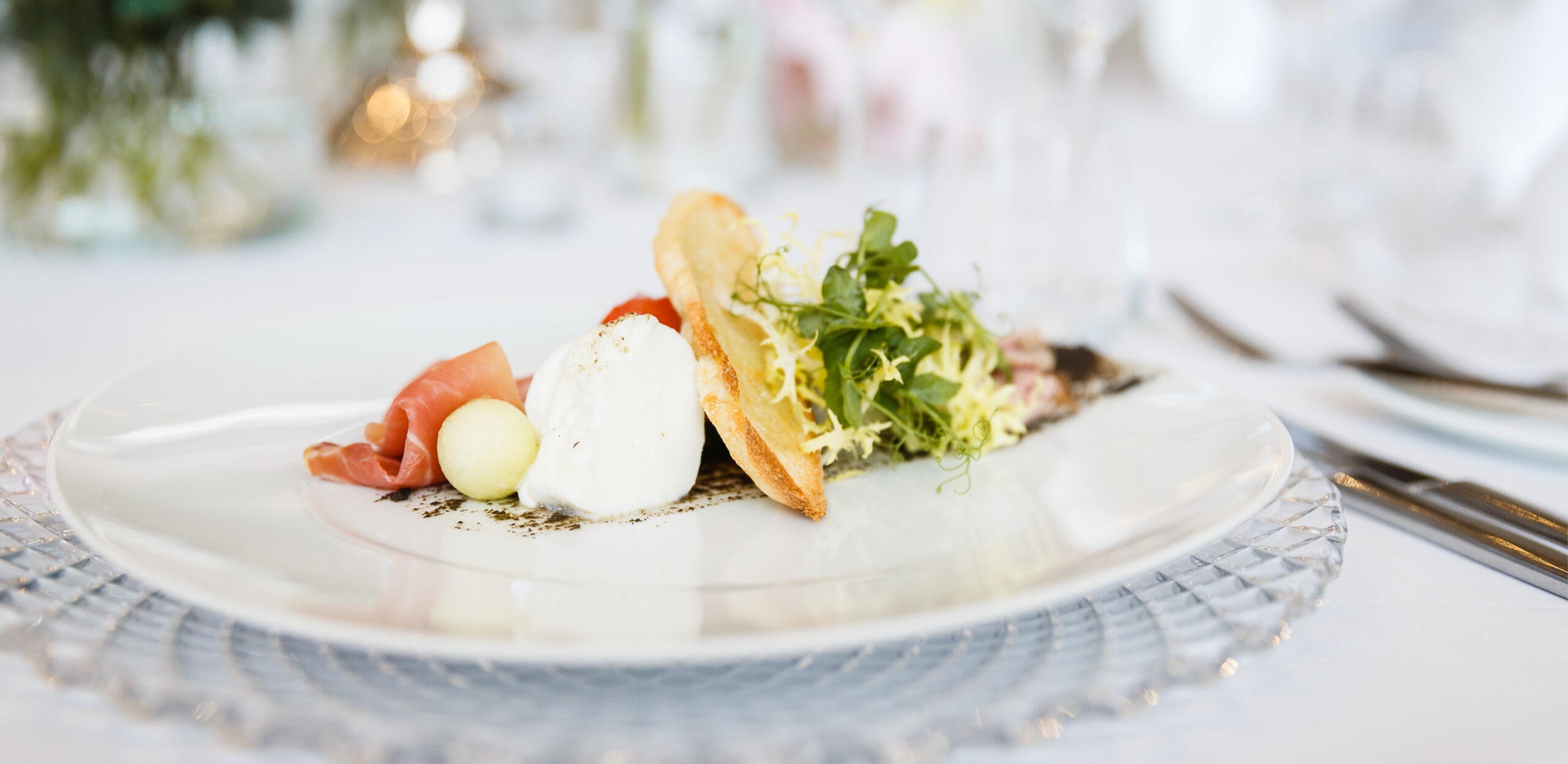 Eine Gourmet-Vorspeise mit Prosciutto, Mozzarella, Melone, Toast und Grünzeug auf einem weißen Teller - perfekt für Catering für Hochzeiten und Feiern.