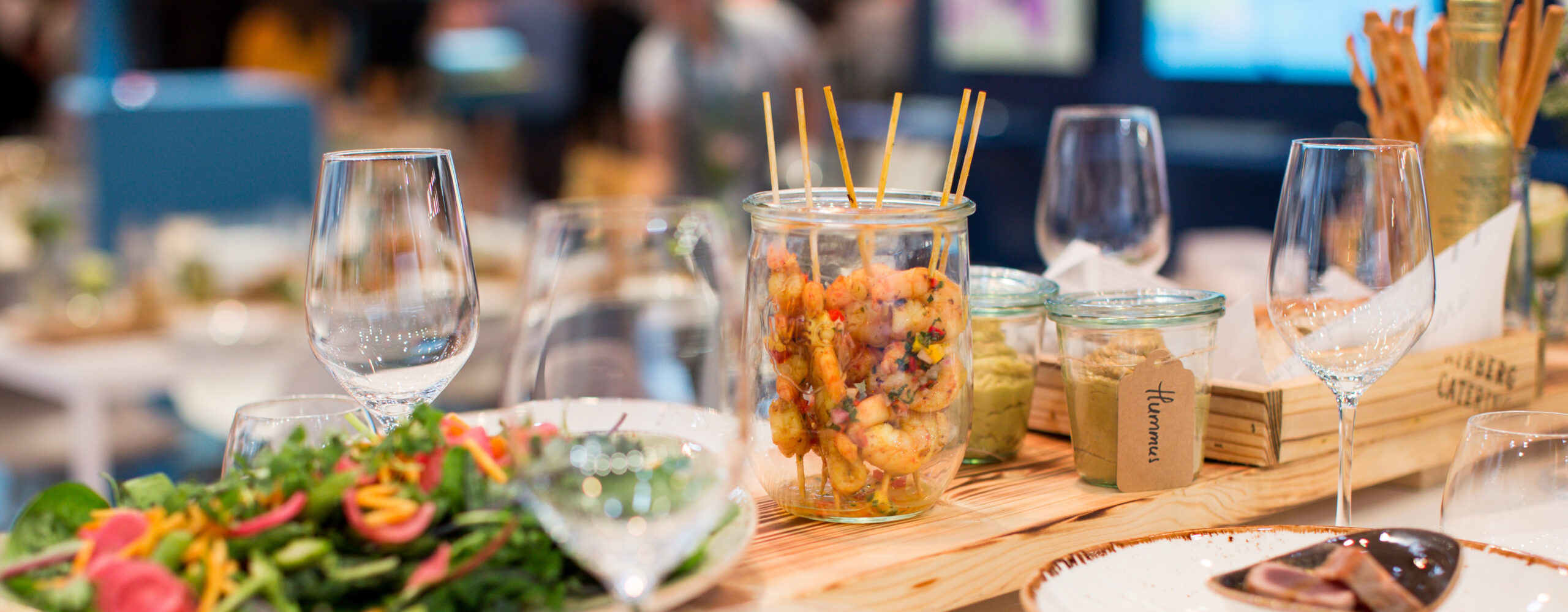 Glasgefäß gefüllt mit aufgespießten Garnelen auf einem Tisch für das Messecatering für Veranstaltungen, mit Salat, leeren Weingläsern und kleinen Gläsern mit Lebensmitteln.