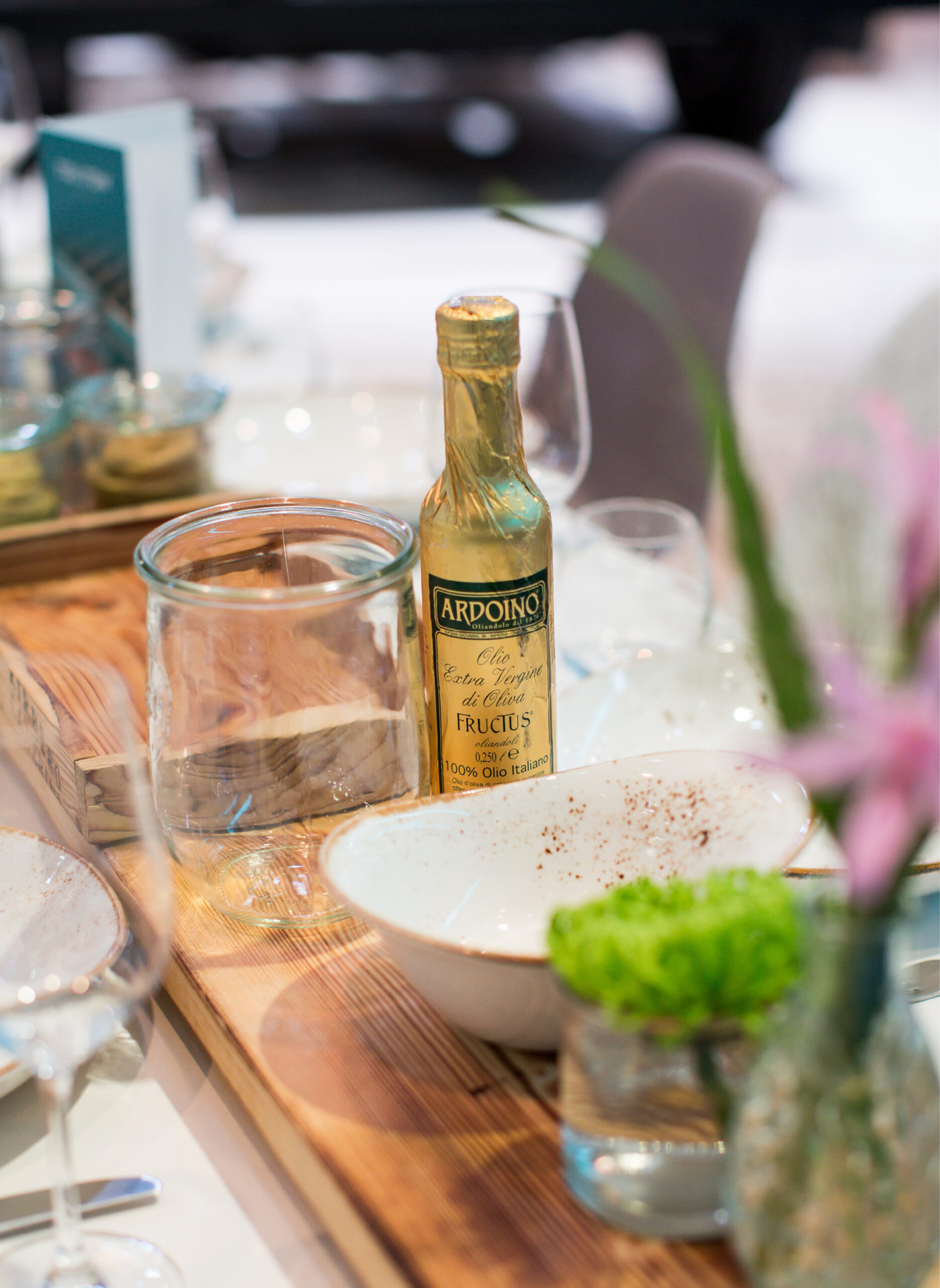 Eine Flasche Olivenöl auf einem Holztablett, umgeben von Gläsern, einer Schale und einer Vase mit Blumen - perfekt für das Messecatering für Veranstaltungen.
