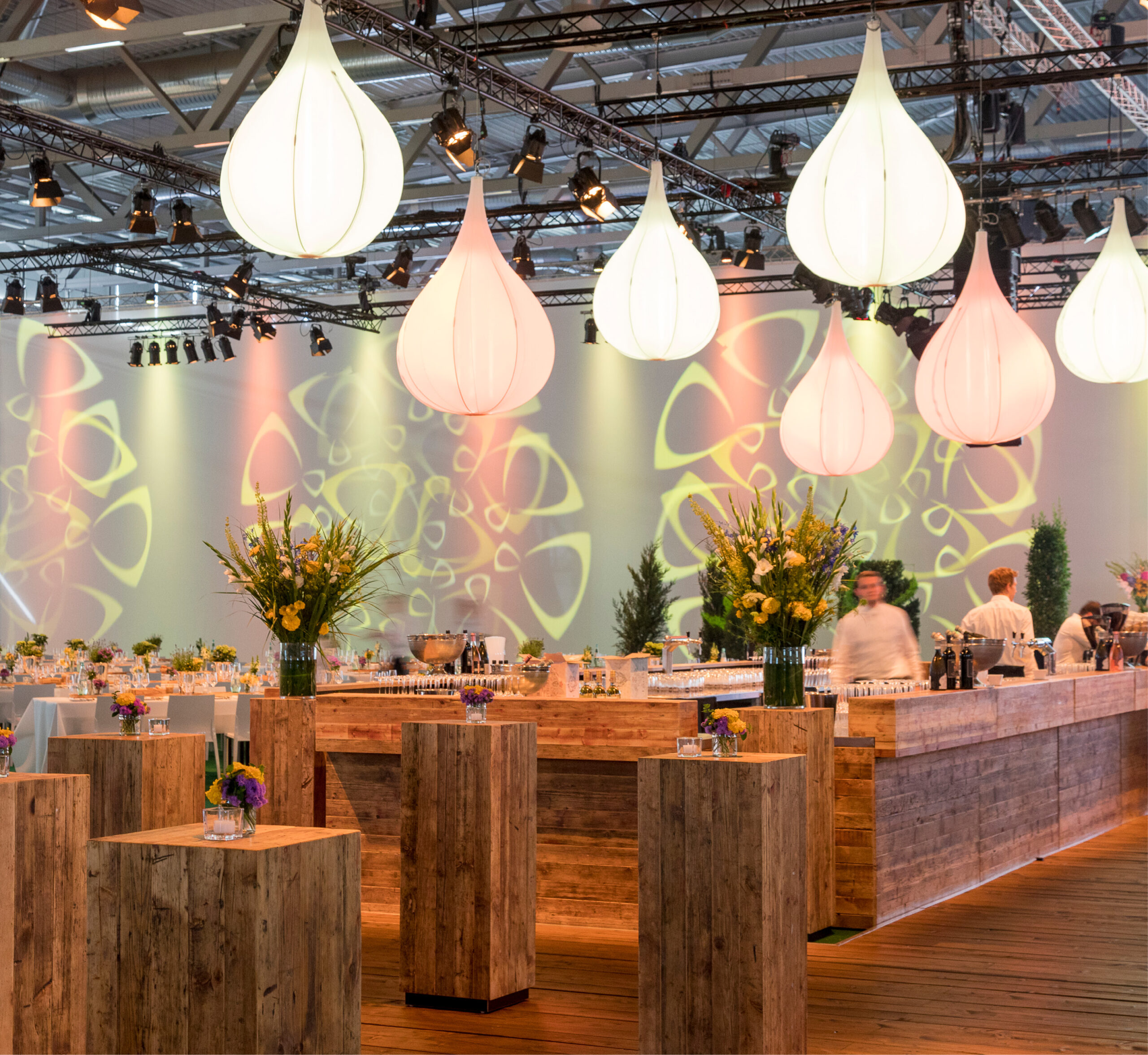 Ein moderner Veranstaltungsraum mit Holztischen, Blumenarrangements und großen tropfenförmigen Hängelampen - perfekt für stilvolles Messecatering für Veranstaltungen.